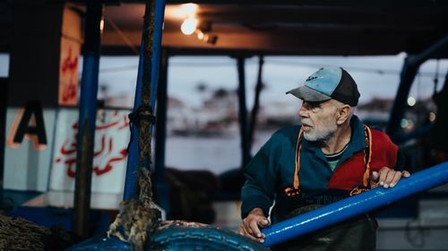 Free An elderly man wearing a cap on a fishing boat during dusk, holding a blue rail. Stock Photo