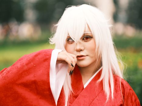 Close-up of a person in red anime costume with white hair outdoors.