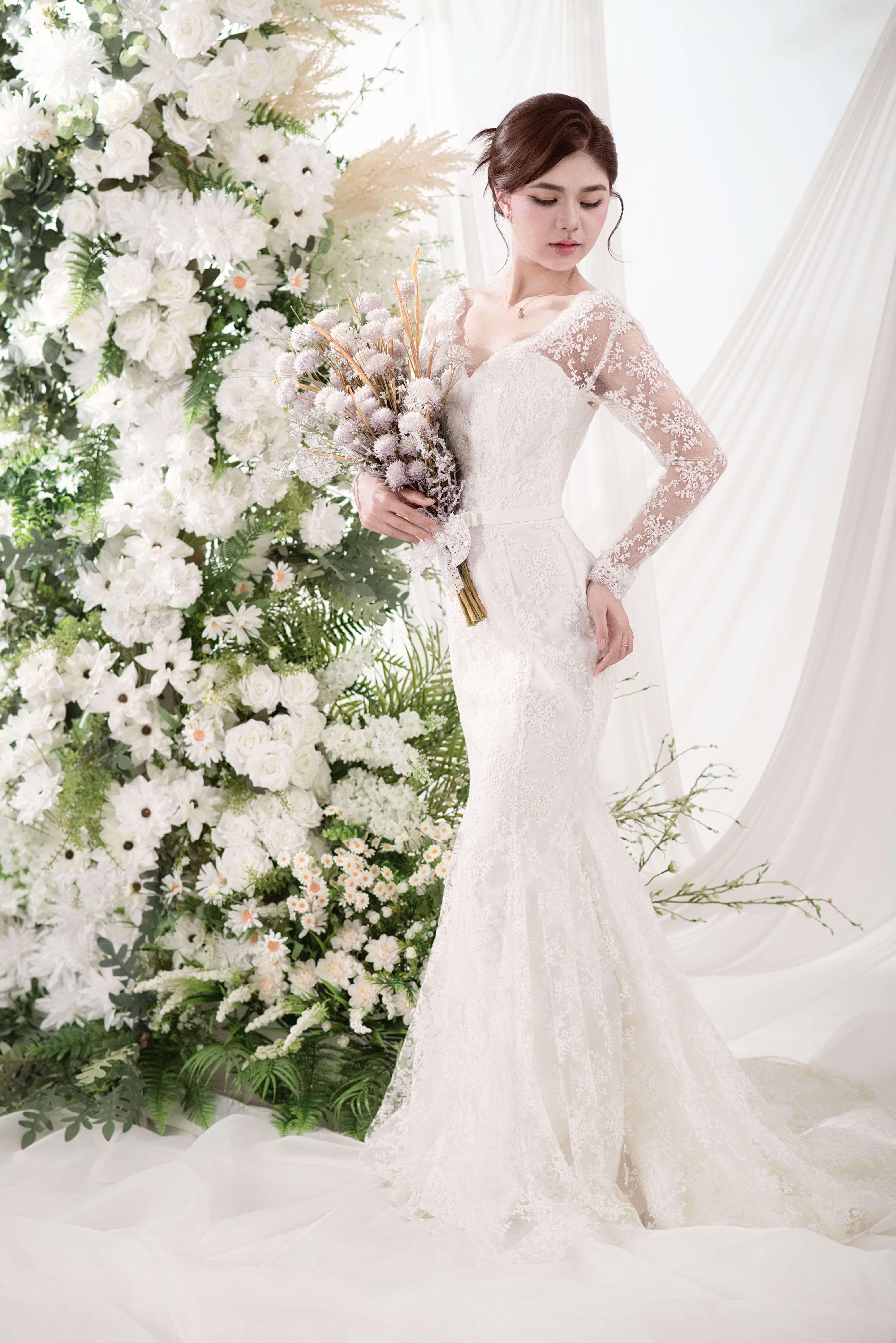 Bride in lace gown holding bouquet in front of lush floral arrangement, exuding elegance and beauty.