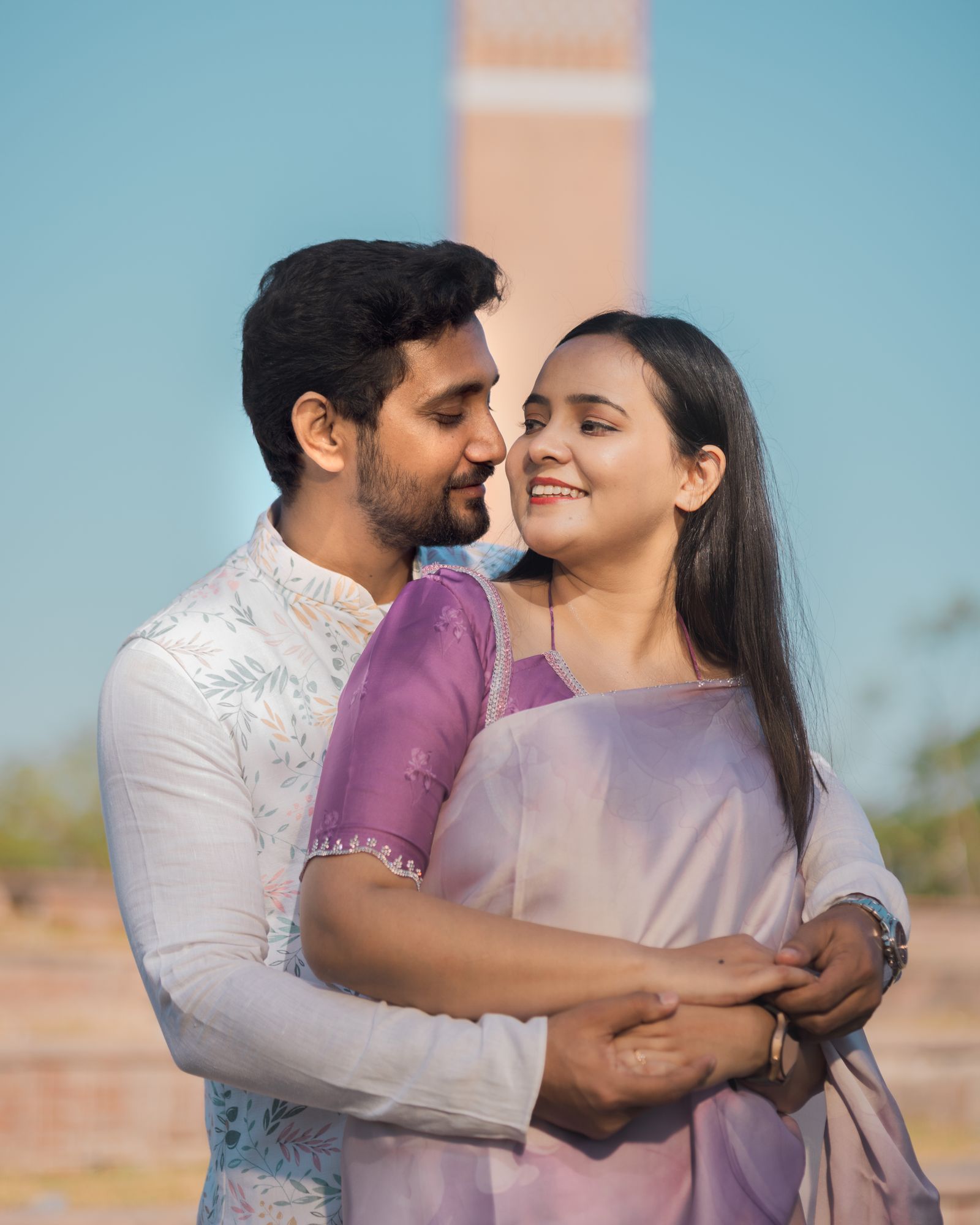 A couple outdoors in mauve and white, his arms around her, both of them laughing.