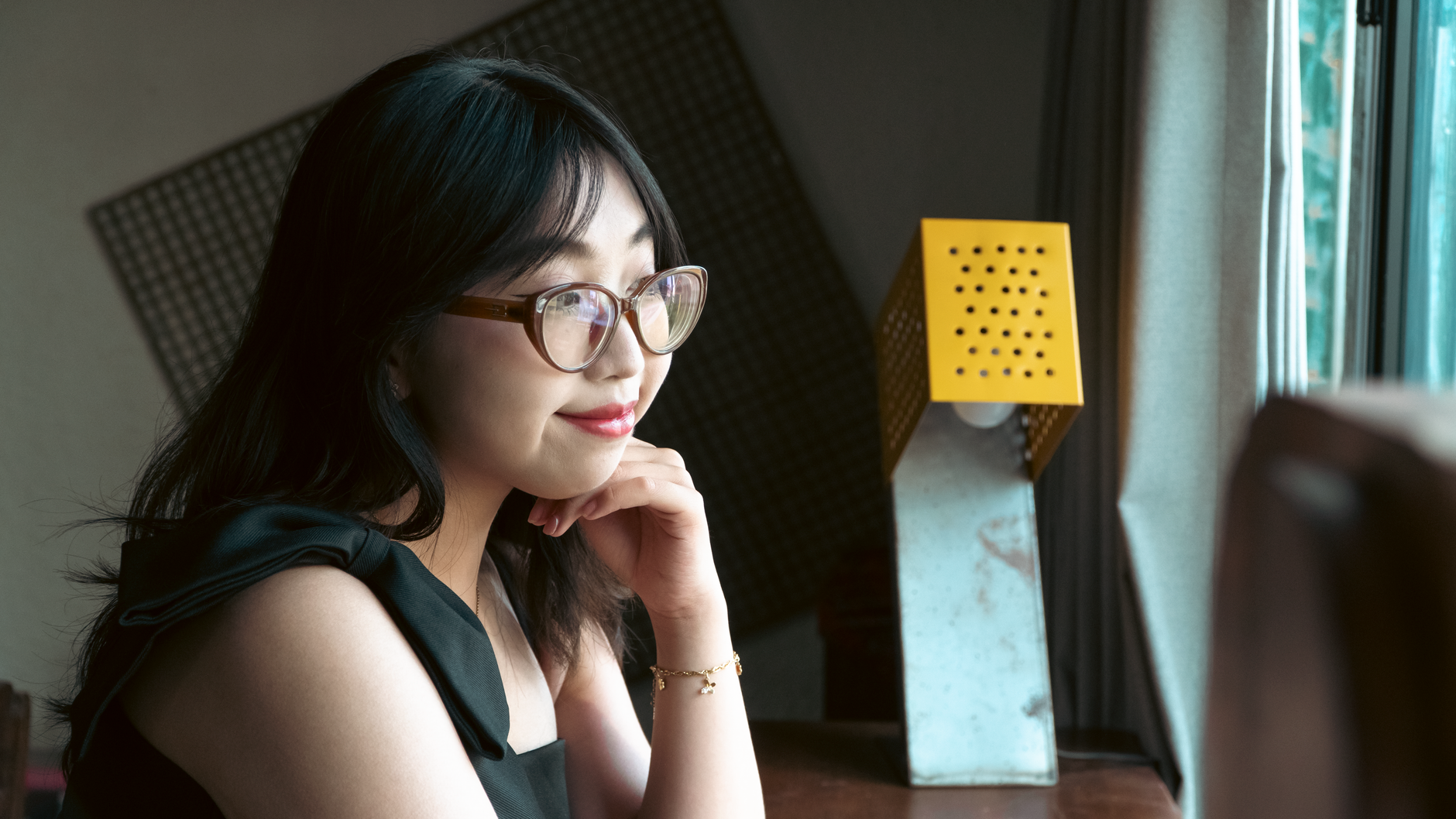 A woman with glasses gazes thoughtfully out a window in a cozy room.