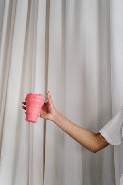 Close-up of a hand holding a pink reusable tumbler against a light curtain background.