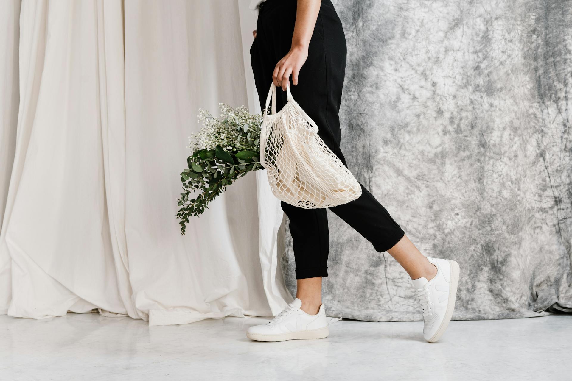 Woman in minimalist outfit holding an eco-friendly mesh bag filled with flowers, showcasing sustainable fashion.