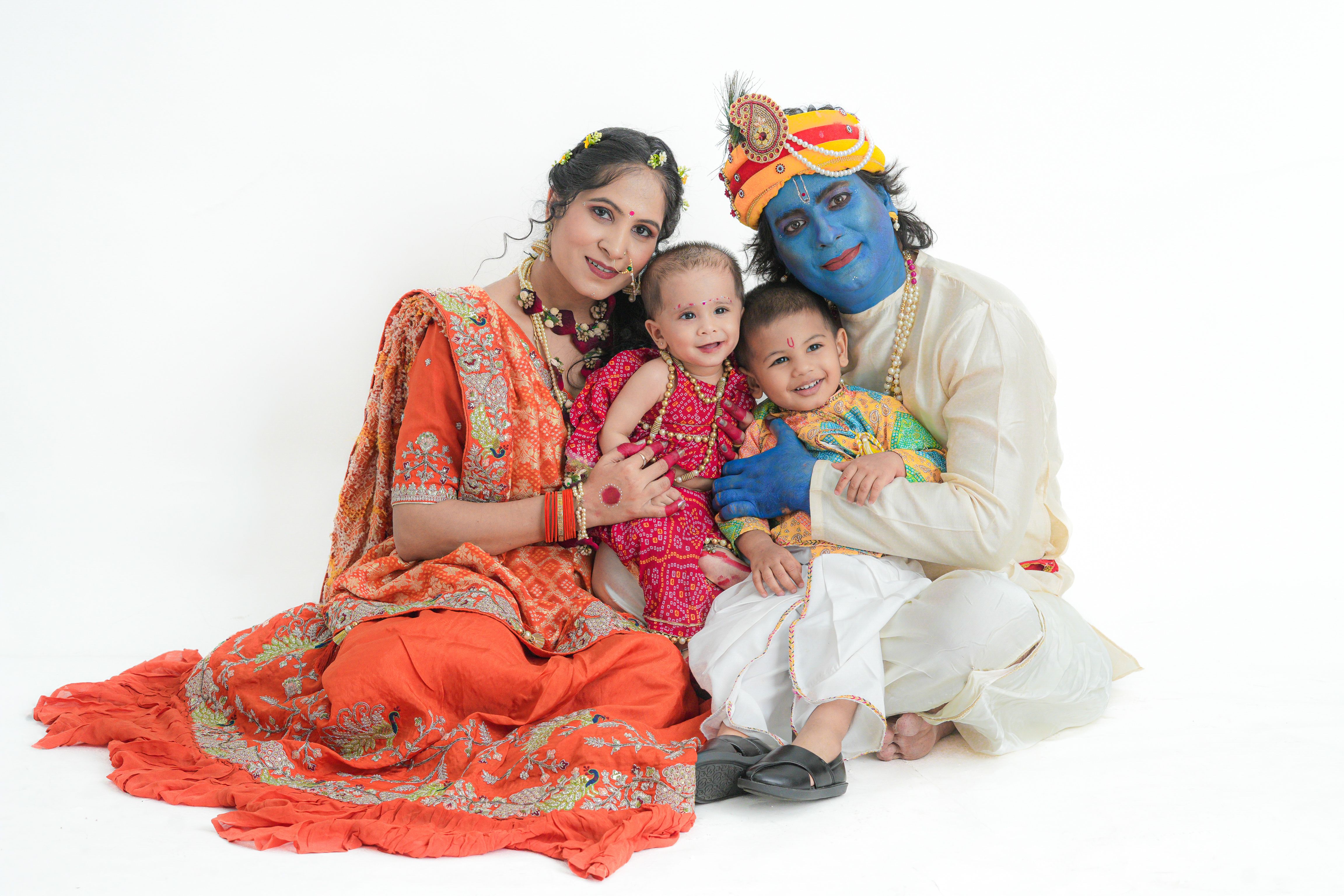 A family of four dressed in traditional attire with one member as Lord Krishna, smiling together against a white background.
