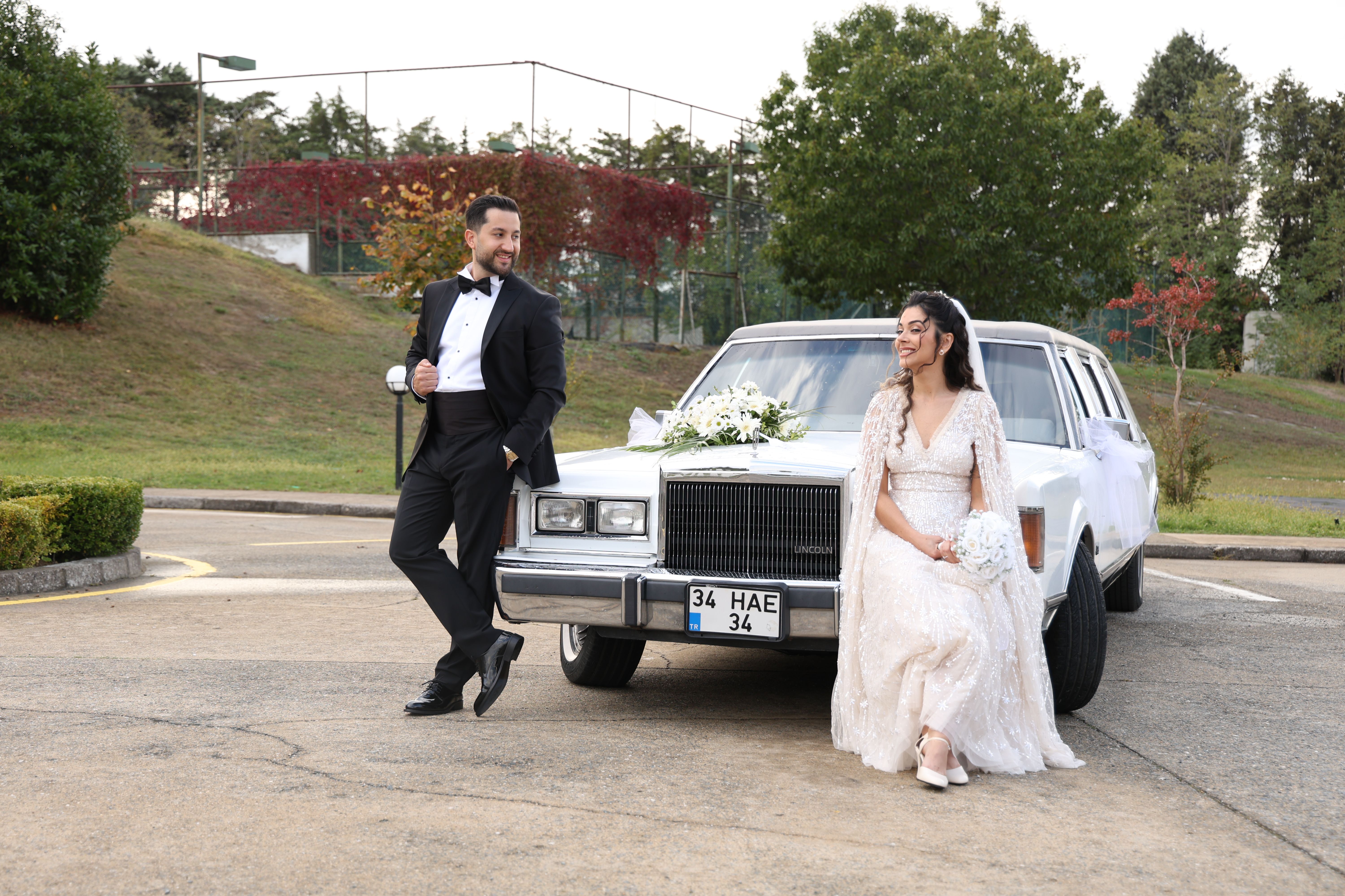 A newlywed couple poses stylishly next to a classic car in an outdoor setting.