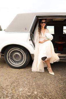 Elegant bride stepping out of a vintage limousine, holding a bouquet, and wearing stylish sunglasses.