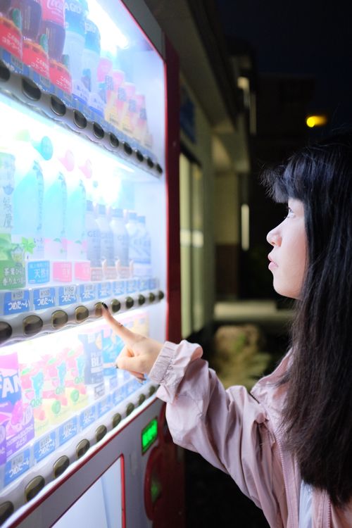 Free A young woman selects a drink from a vending machine, illuminated at night in an urban setting. Stock Photo