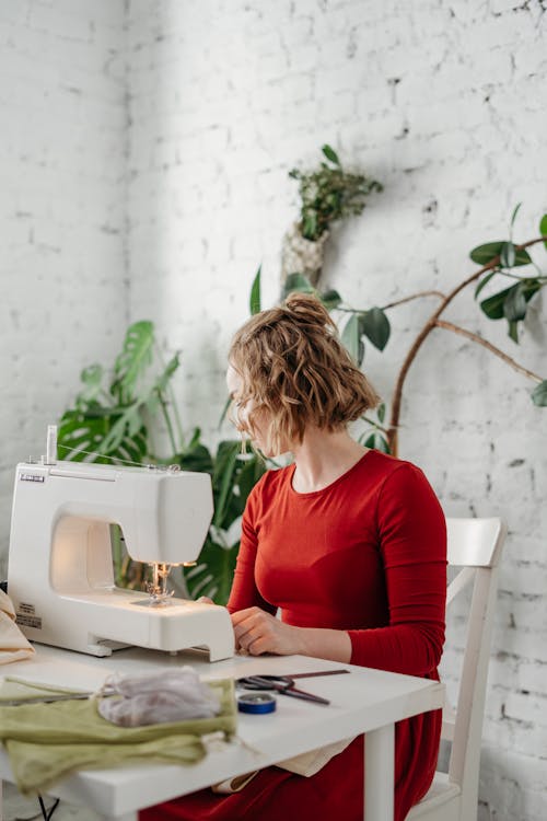 Woman in Red Long Sleeve Dress Sitting on Chair Using Sewing Machine