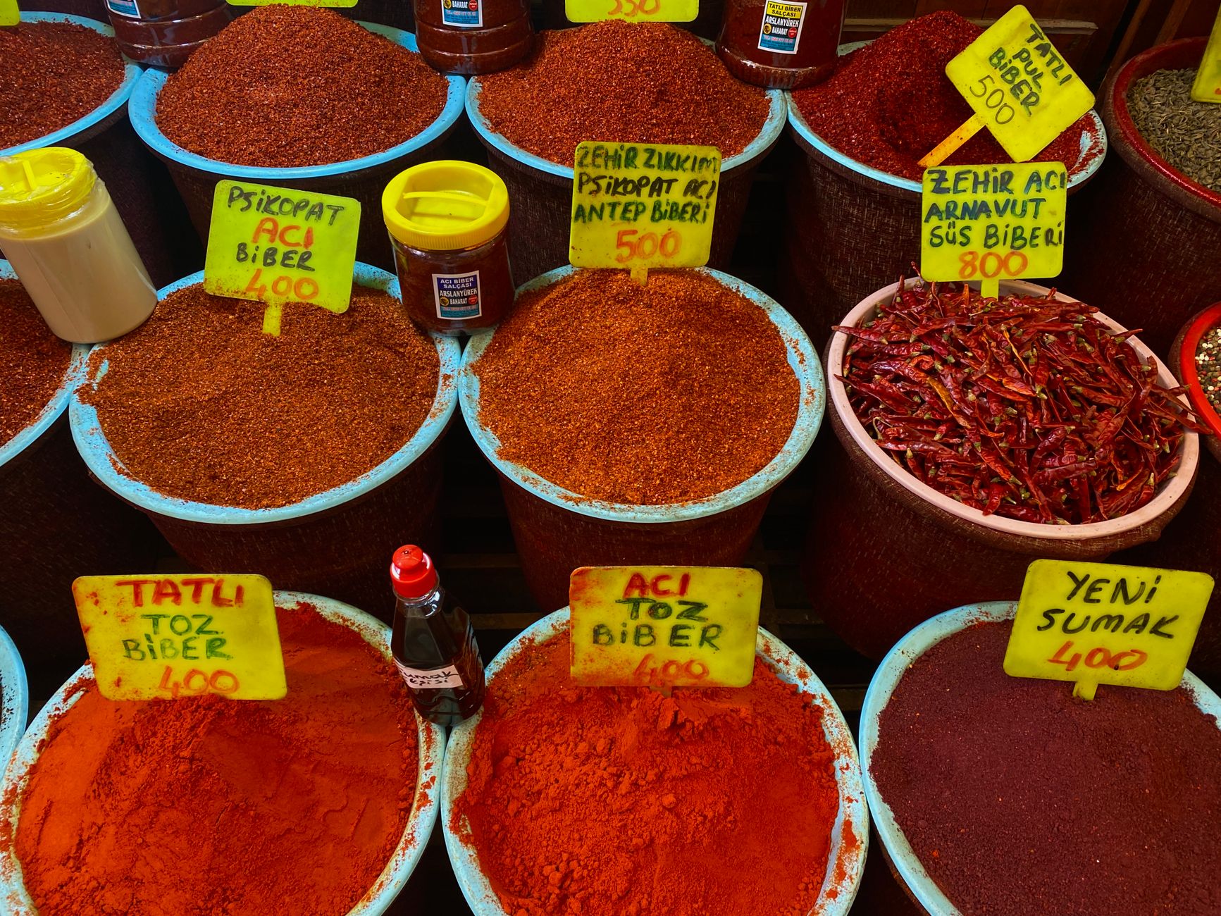 Colorful array of Middle Eastern spices and chile pastes in small ceramic bowls, featuring dark red harissa, burgundy urfa biber flakes, and bright Aleppo pepper on a rustic wooden table
