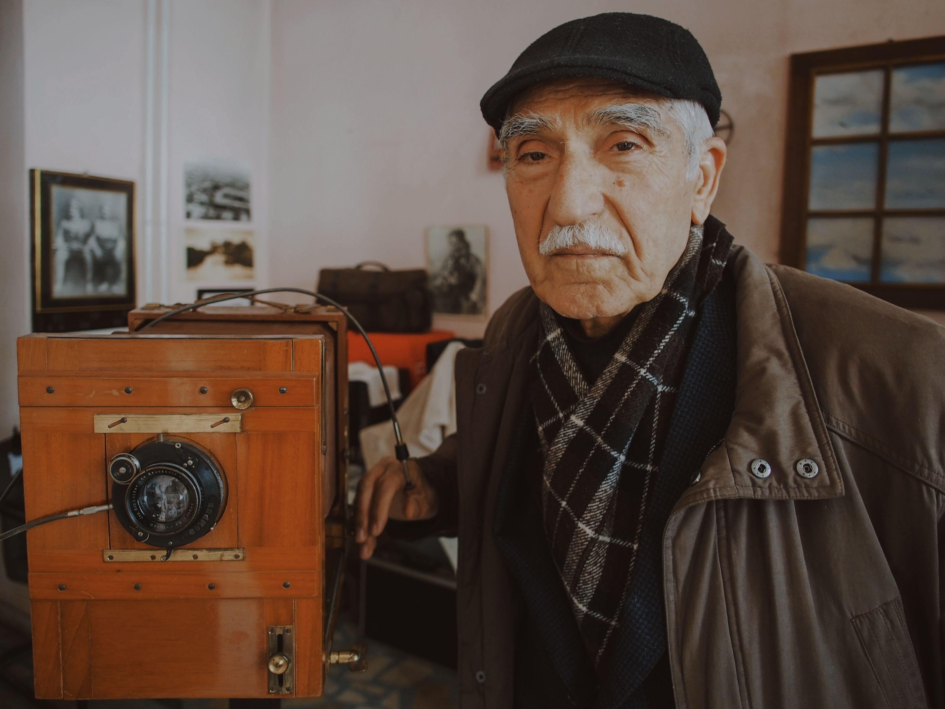 Elderly man with a cap poses beside an antique camera, showcasing vintage photography equipment.