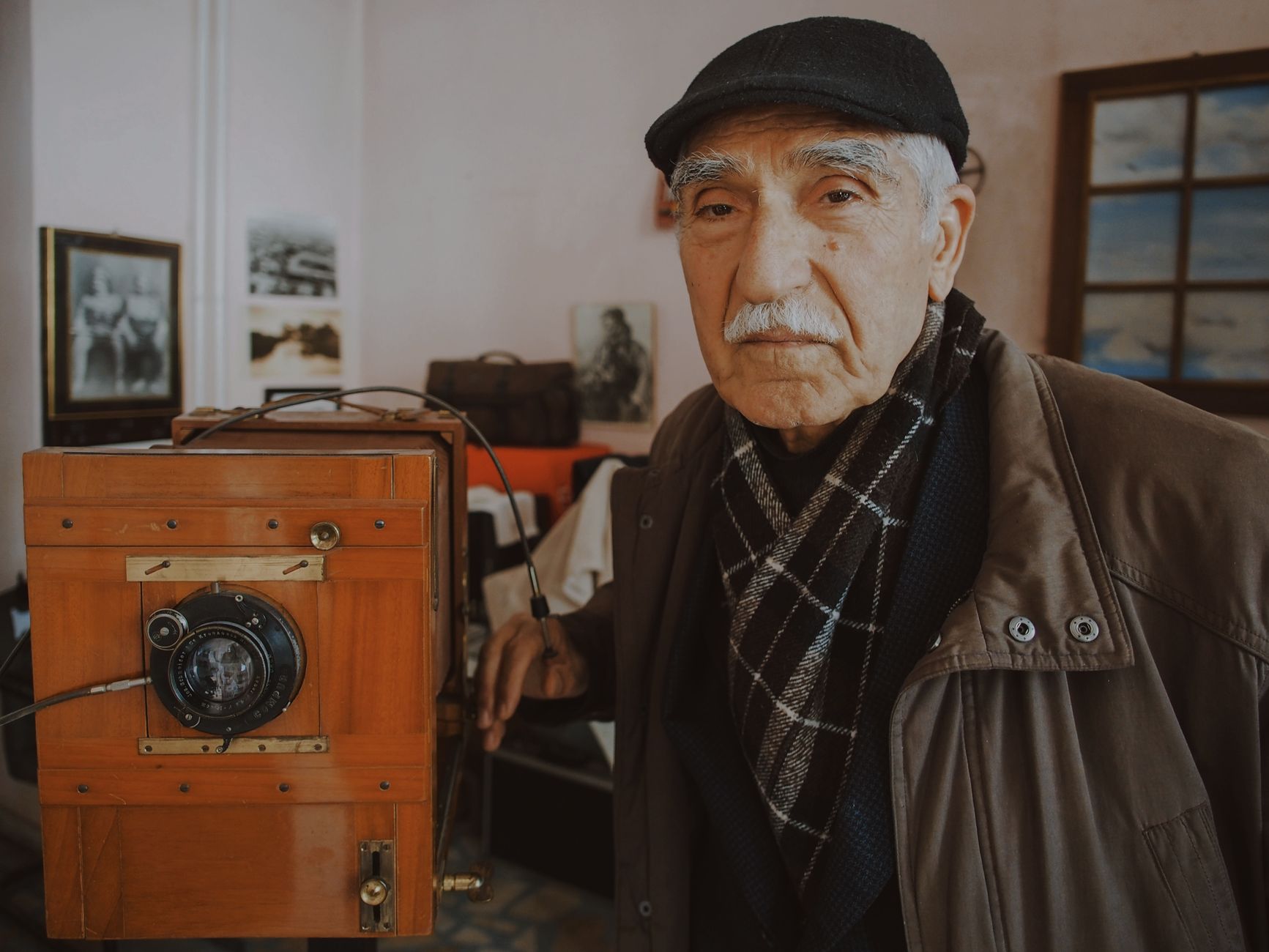 Elderly man with a cap poses beside an antique camera, showcasing vintage photography equipment.