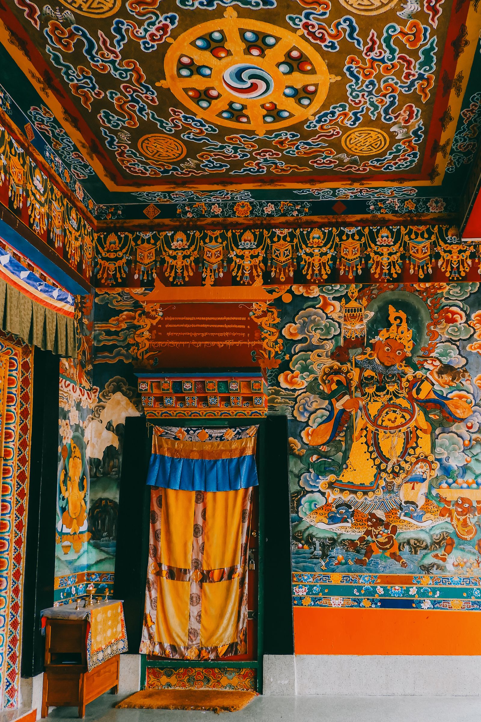 The porch of Rumtek Monastery, a painted ceiling mandala above a guardian king mural and a curtained door to the hall