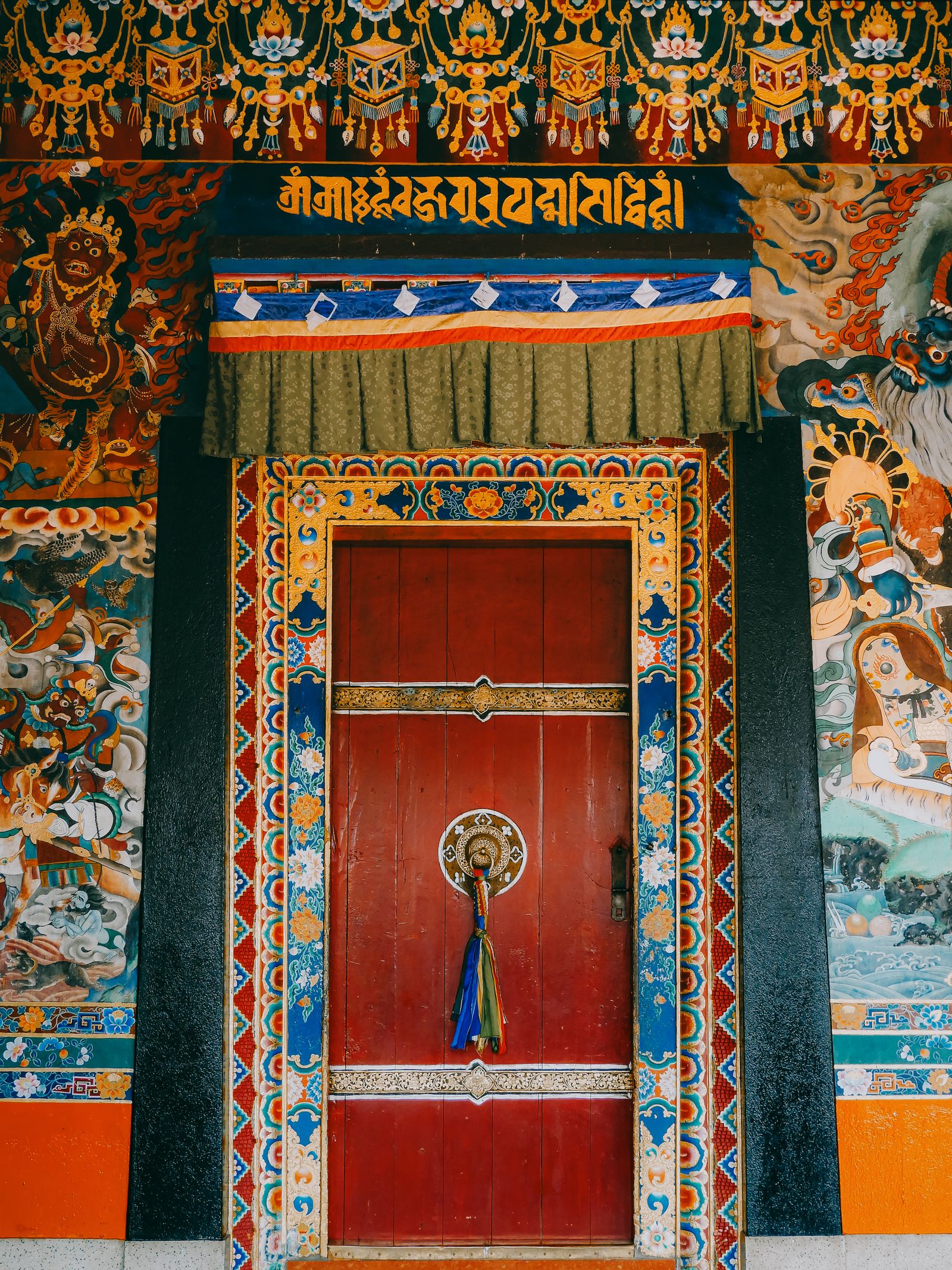 A red door hung with a silk tassel on the porch of Rumtek Monastery, framed by painted guardian kings and Tibetan script