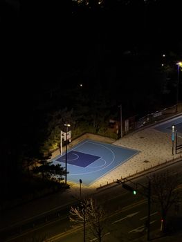Dramatic aerial view of a lit-up basketball court at night, surrounded by urban elements.