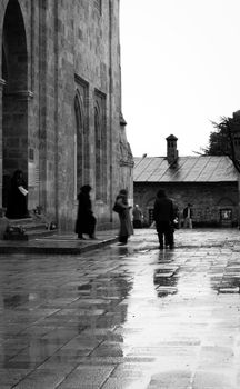 Black and white photo of people walking in a rain-soaked courtyard beside historic architecture.