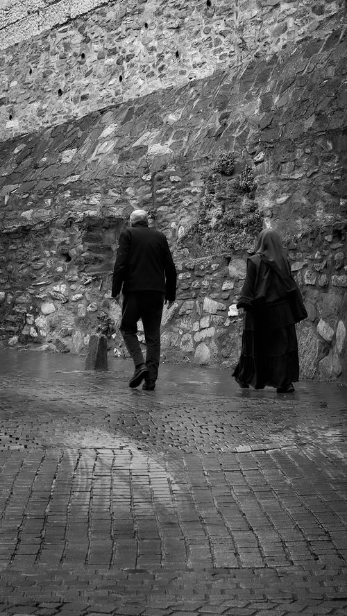 Free Black and white image of two people walking along a wet cobblestone path by a stone wall. Stock Photo