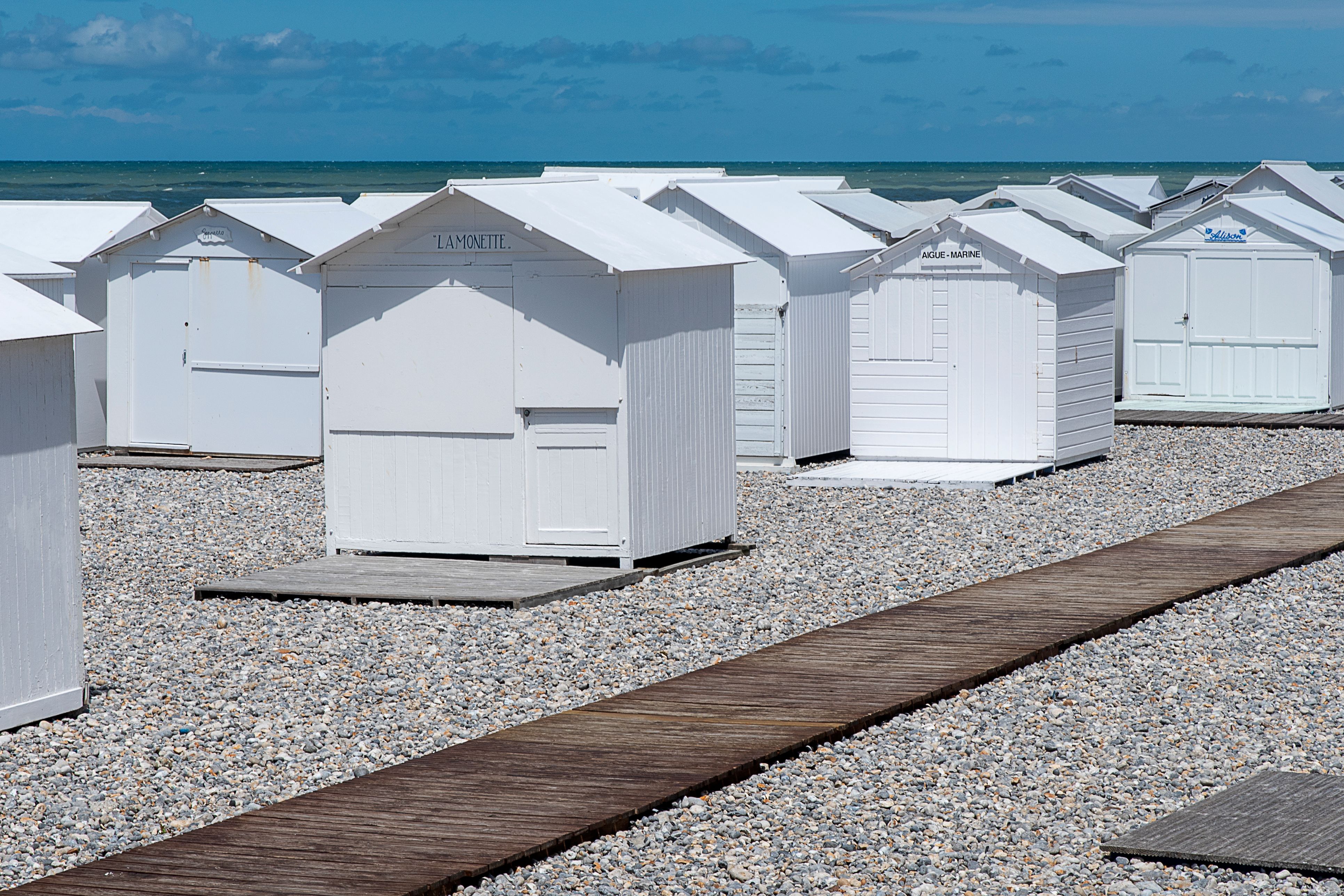 White beach huts line a pebble beach in Mers-les-Bains, France, under a clear blue sky.