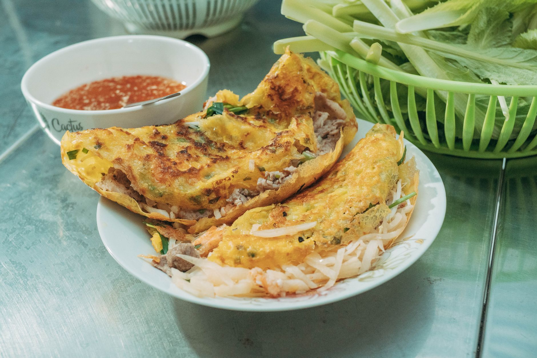 Crispy Vietnamese Bánh Xèo served with fresh herbs and traditional dipping sauce on a metal table.