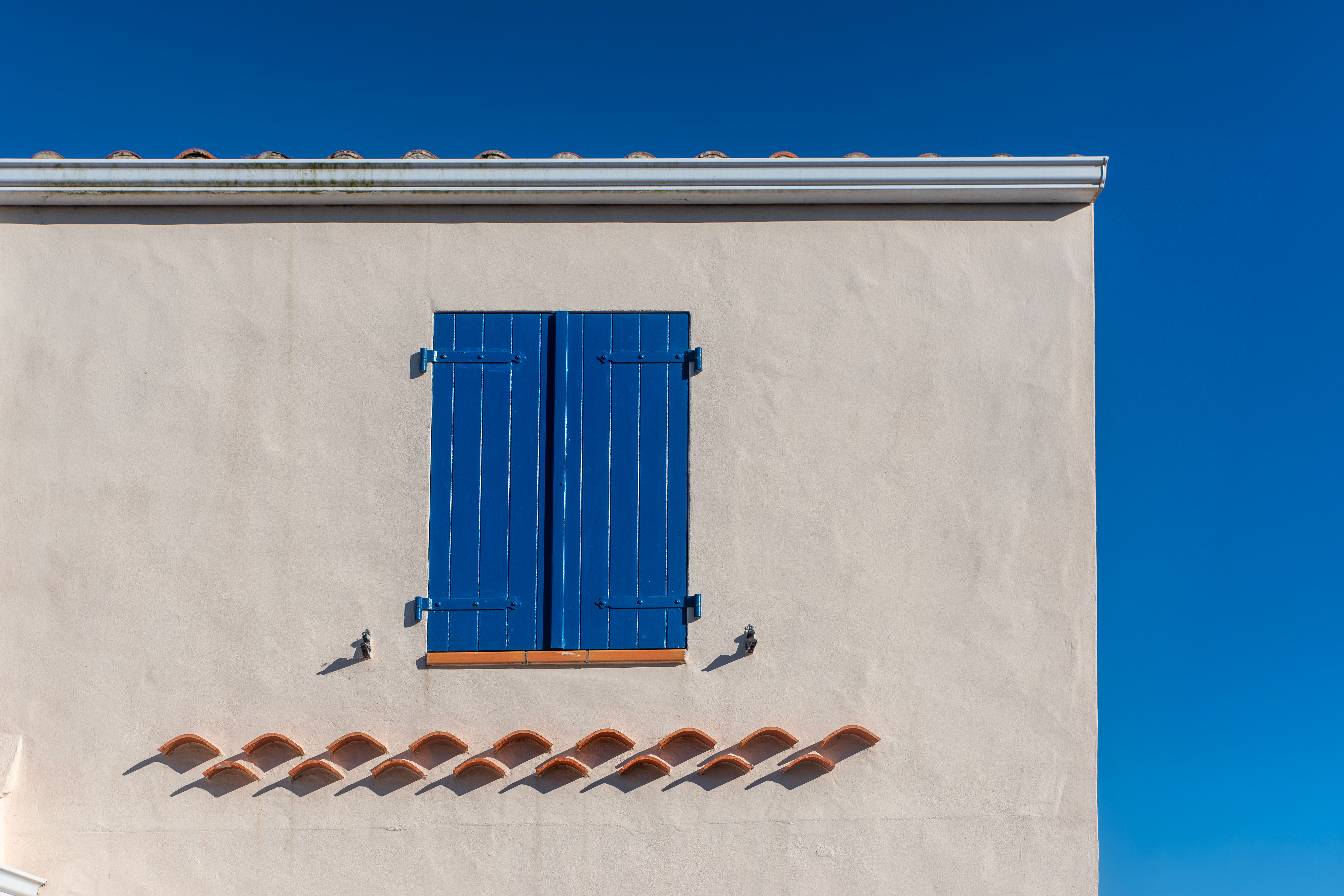 Photo minimaliste d'une fenêtre à volets bleus sur un mur beige, située à Noirmoutier-en-l'Île, en France.