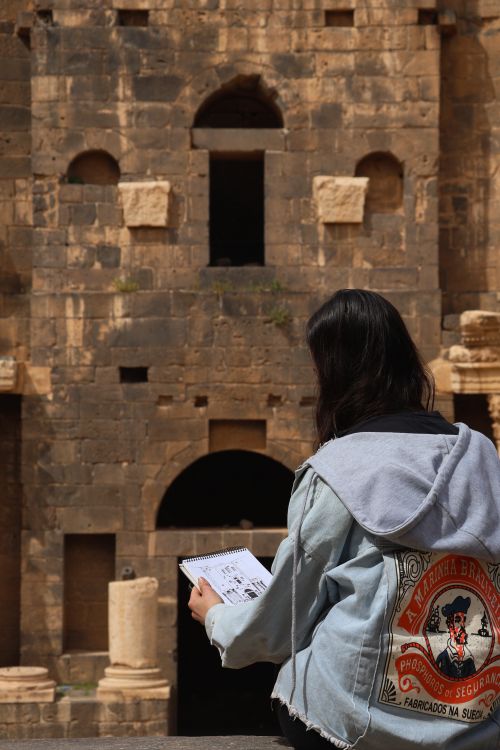 Free Back view of a woman sketching an ancient stone structure outdoors, showcasing artistic creativity. Stock Photo