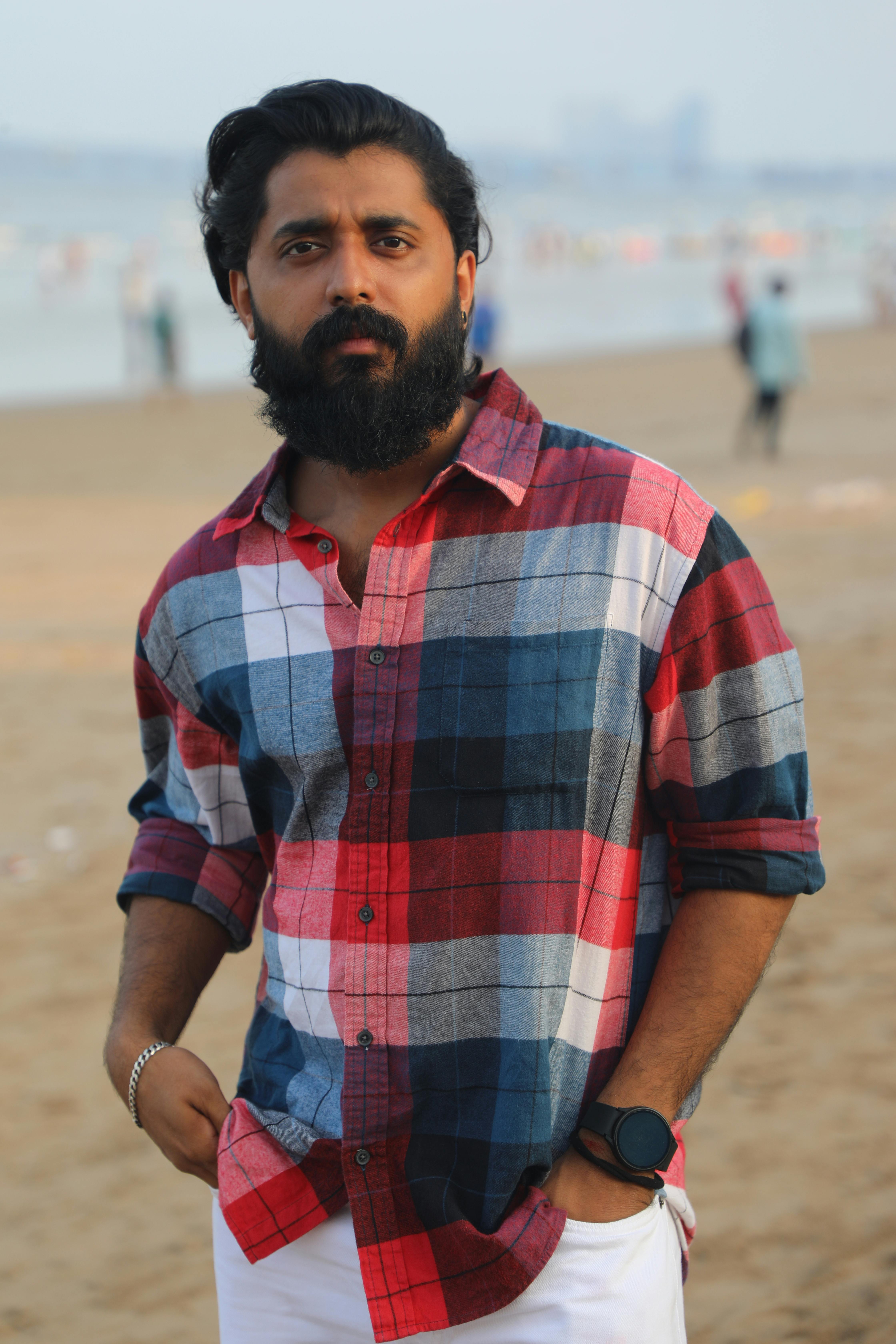 A man in a checkered shirt stands on a sandy beach, exuding a relaxed vibe.