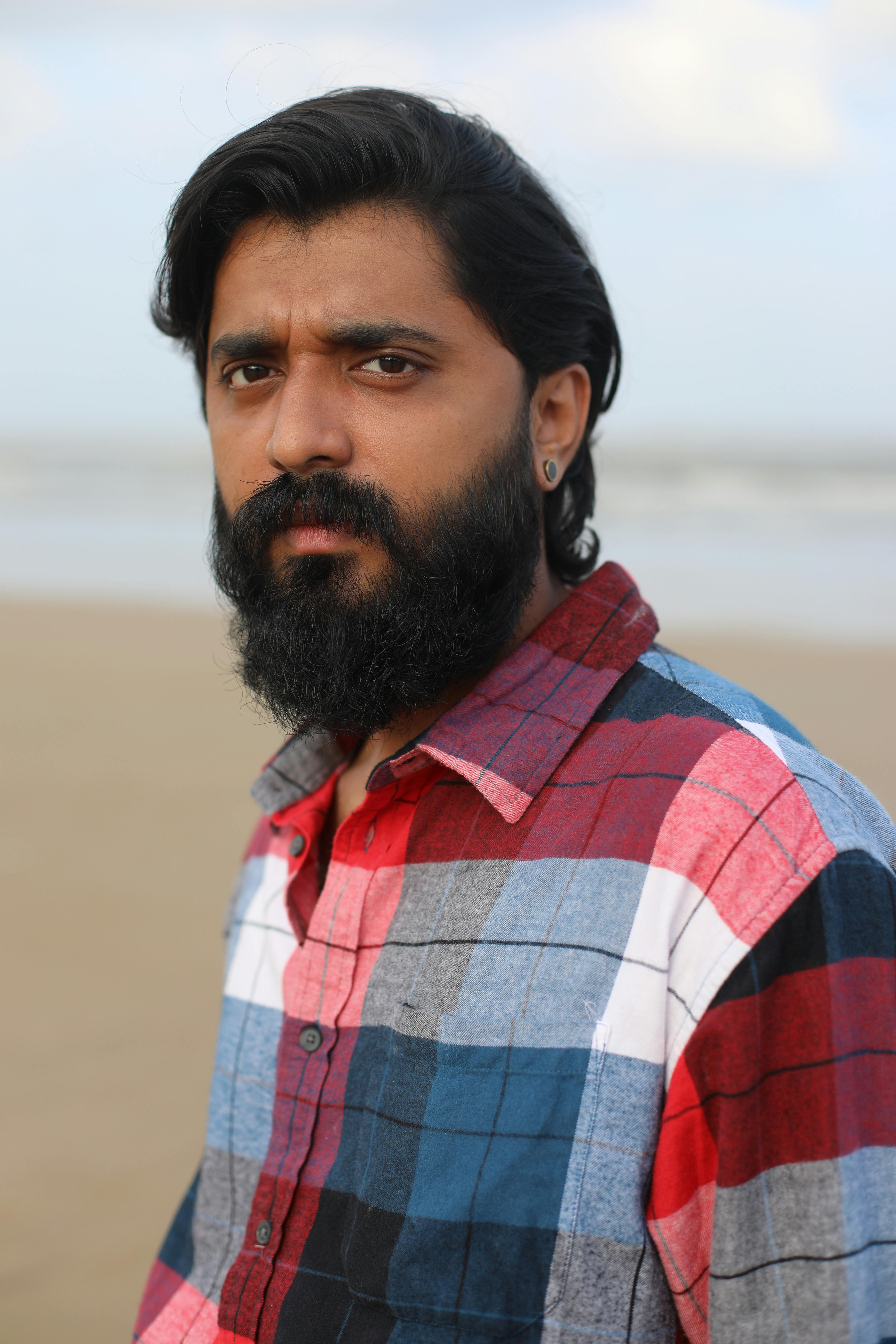 Portrait of a bearded man wearing a plaid shirt standing on a beach during the day.