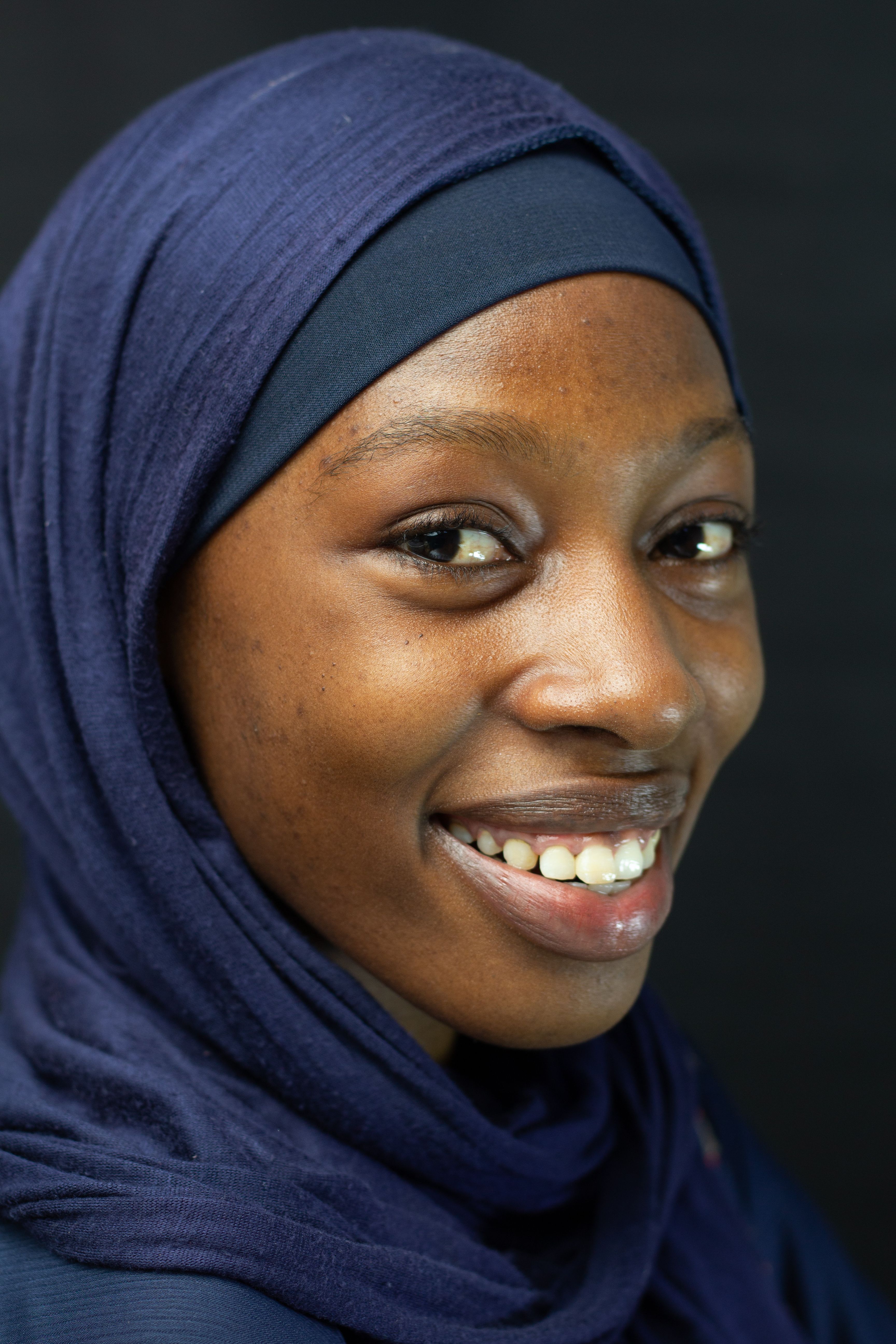 Close-up portrait of a smiling woman wearing a blue hijab with a black background.