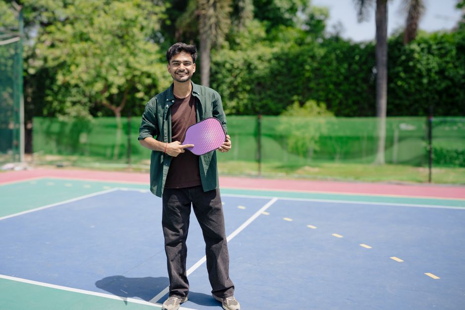 A young man stands on a pickleball court holding a paddle, enjoying a sunny day outdoors.