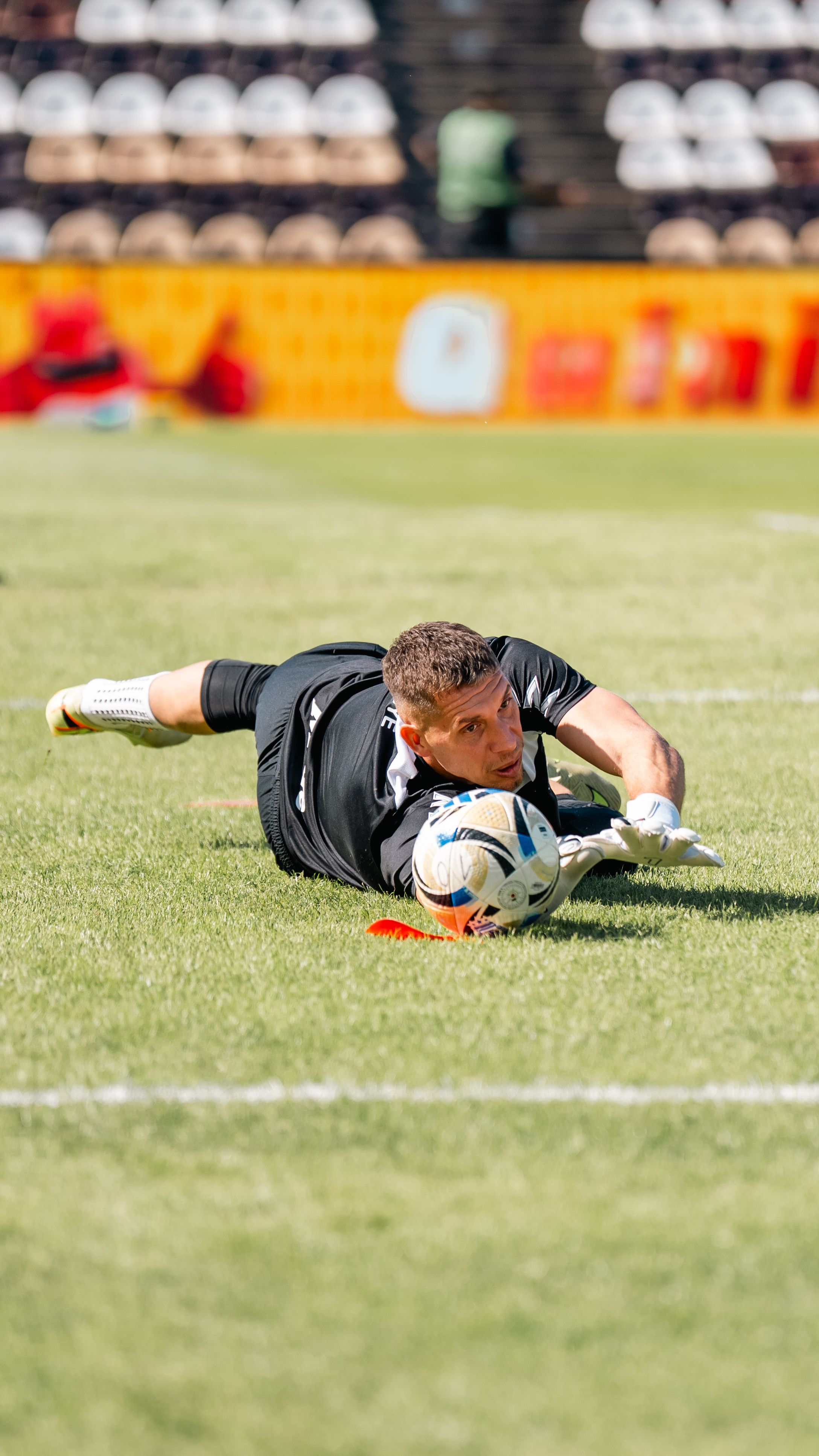Professional goalkeeper dives to save a soccer ball during a sunny outdoor match on a grass field.