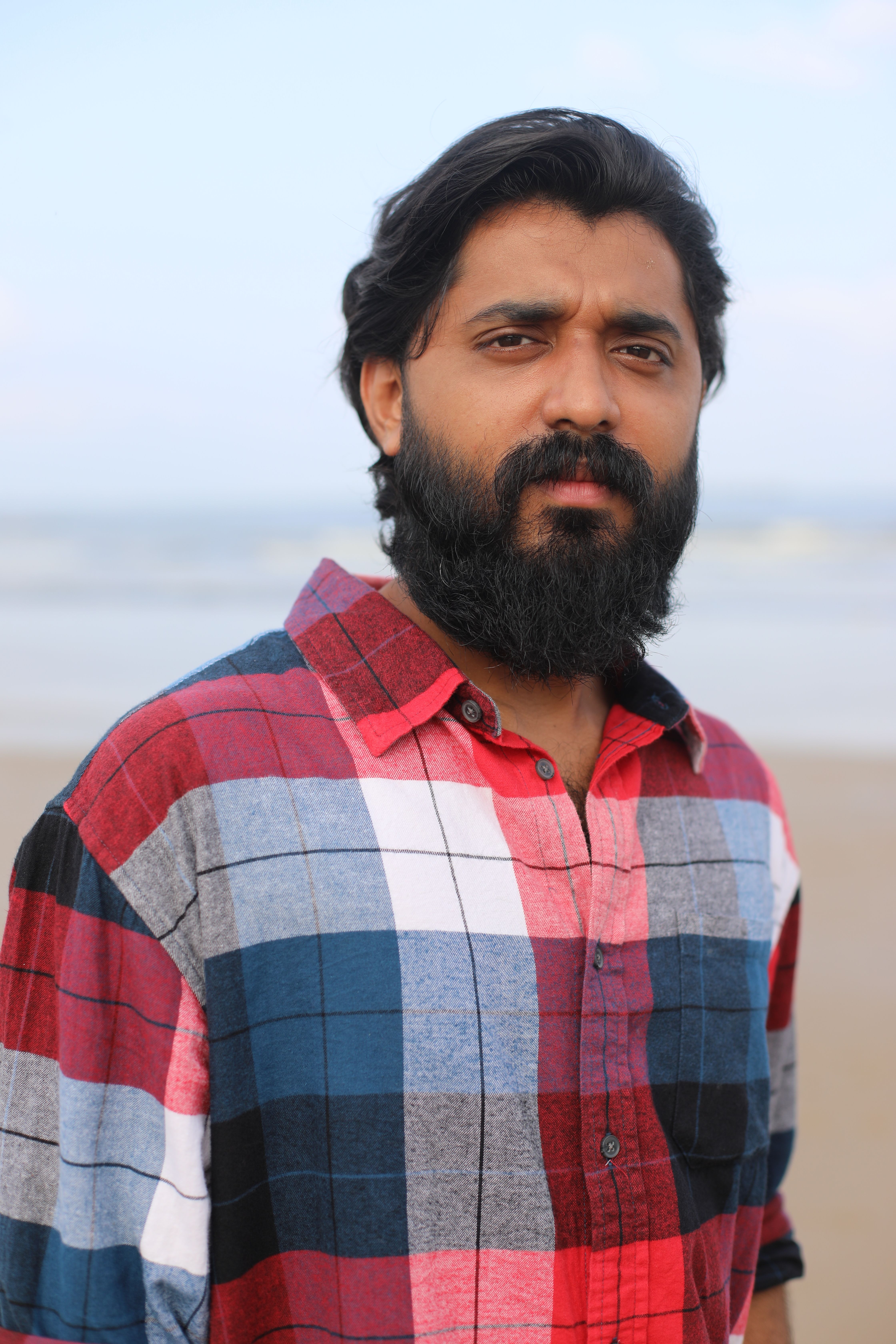 Bearded man in checked shirt poses by the beach, capturing a relaxed vibe.