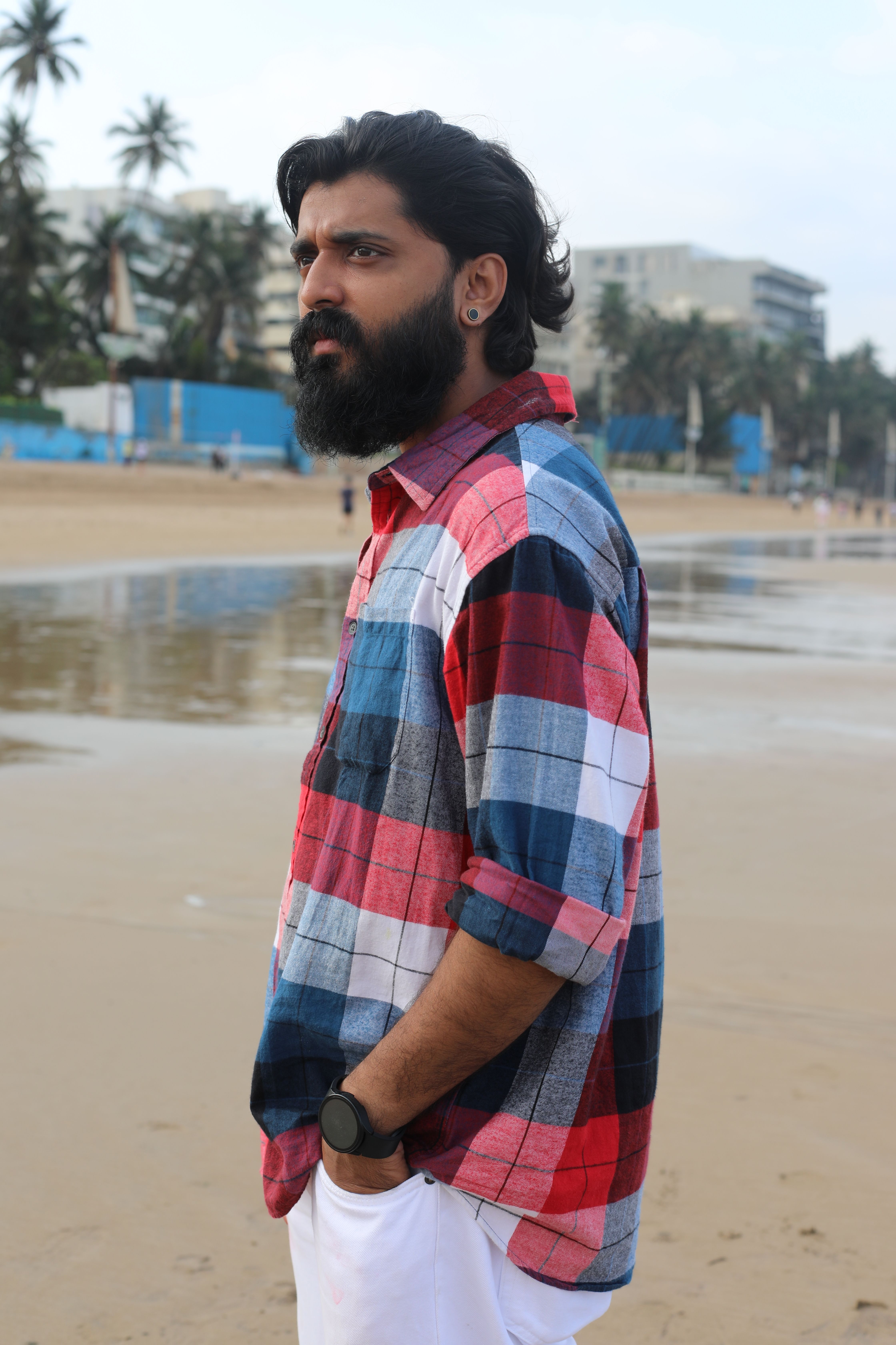 Bearded man in a checkered shirt gazing thoughtfully on a sandy beach.