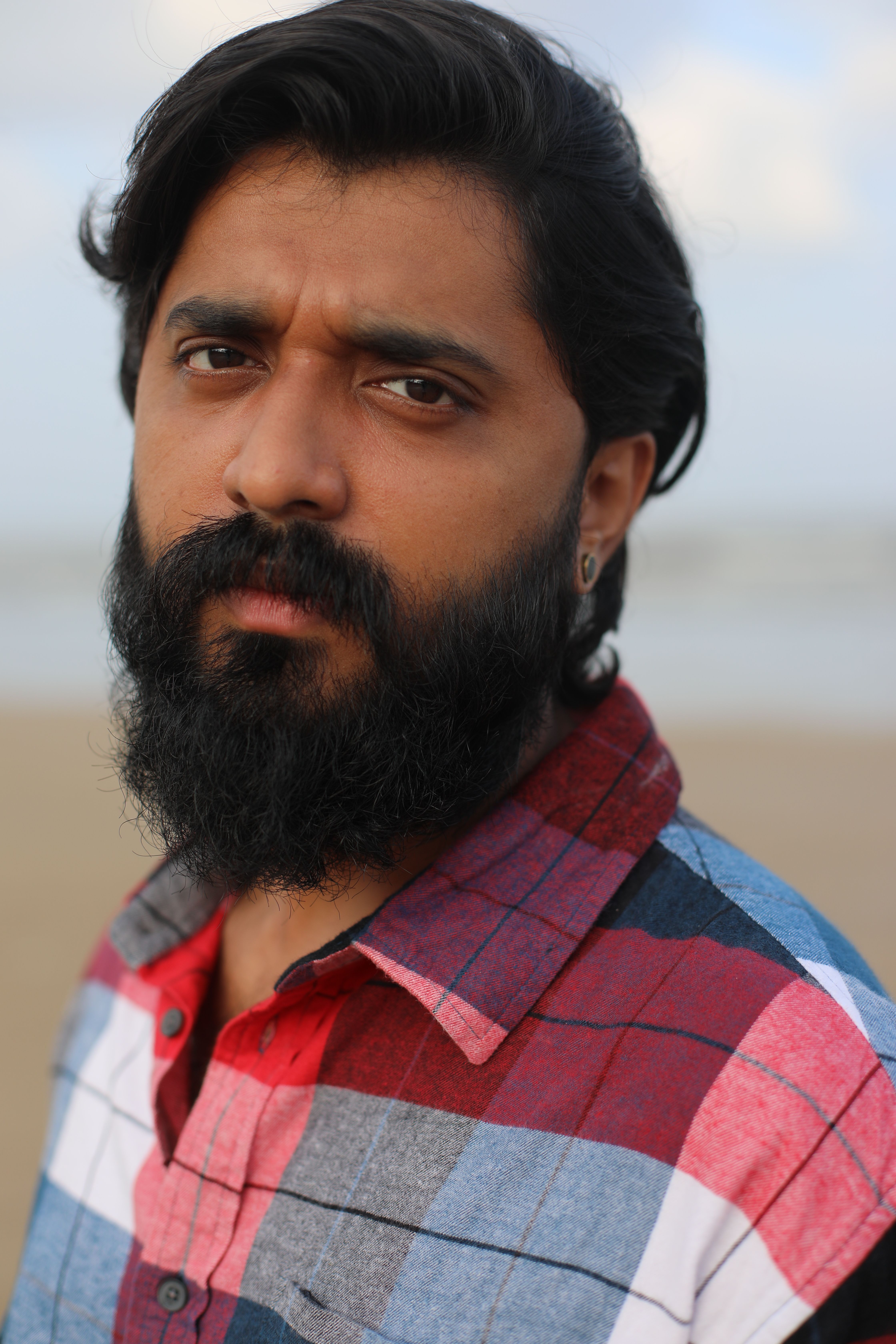 Close-up portrait of a man with a beard standing on a beach, wearing a checked shirt.