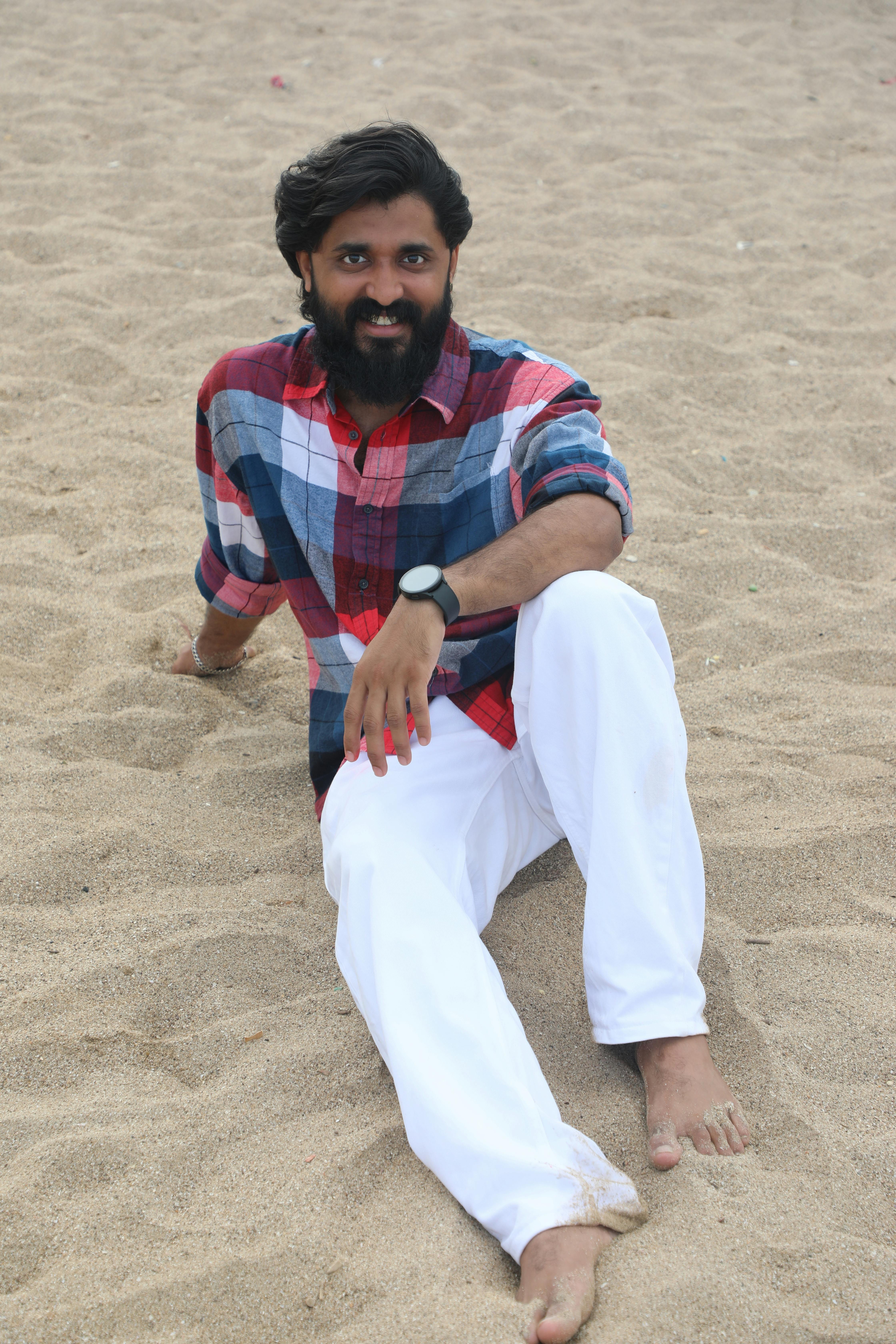 Casual portrait of a man sitting on a sandy beach in a plaid shirt and white trousers.