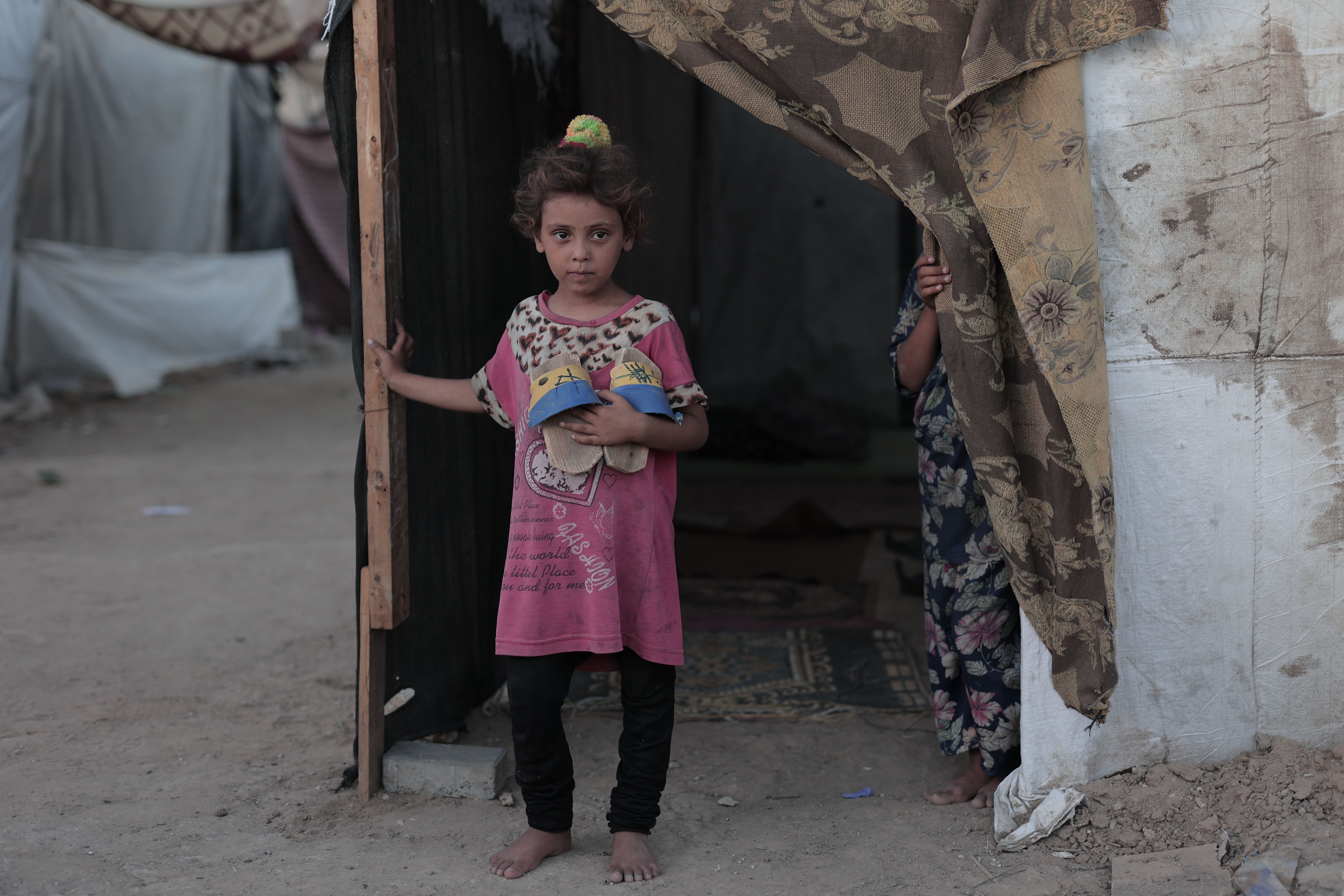 A young child holding belongings at the entrance of a tent in a refugee camp, capturing a poignant moment.