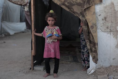 Free A young child holding belongings at the entrance of a tent in a refugee camp, capturing a poignant moment. Stock Photo