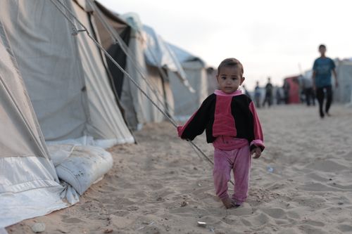 Free A young child walks through a sandy refugee camp, holding a tent rope. Stock Photo