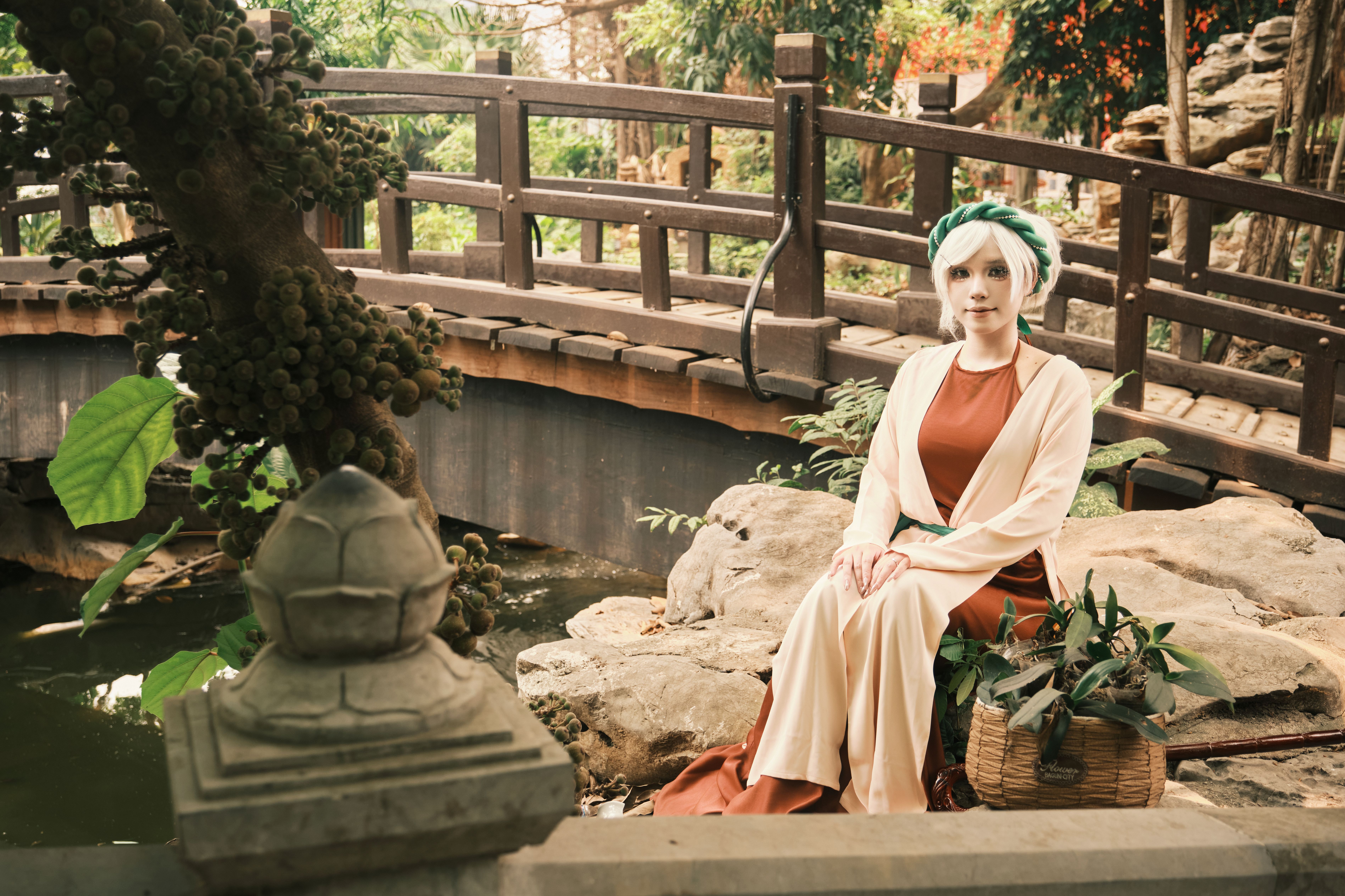 A cosplayer in traditional attire poses on a scenic Vietnamese garden bridge, surrounded by lush greenery.
