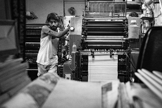 Industrial print shop scene with a worker operating a vintage printing press in black and white.