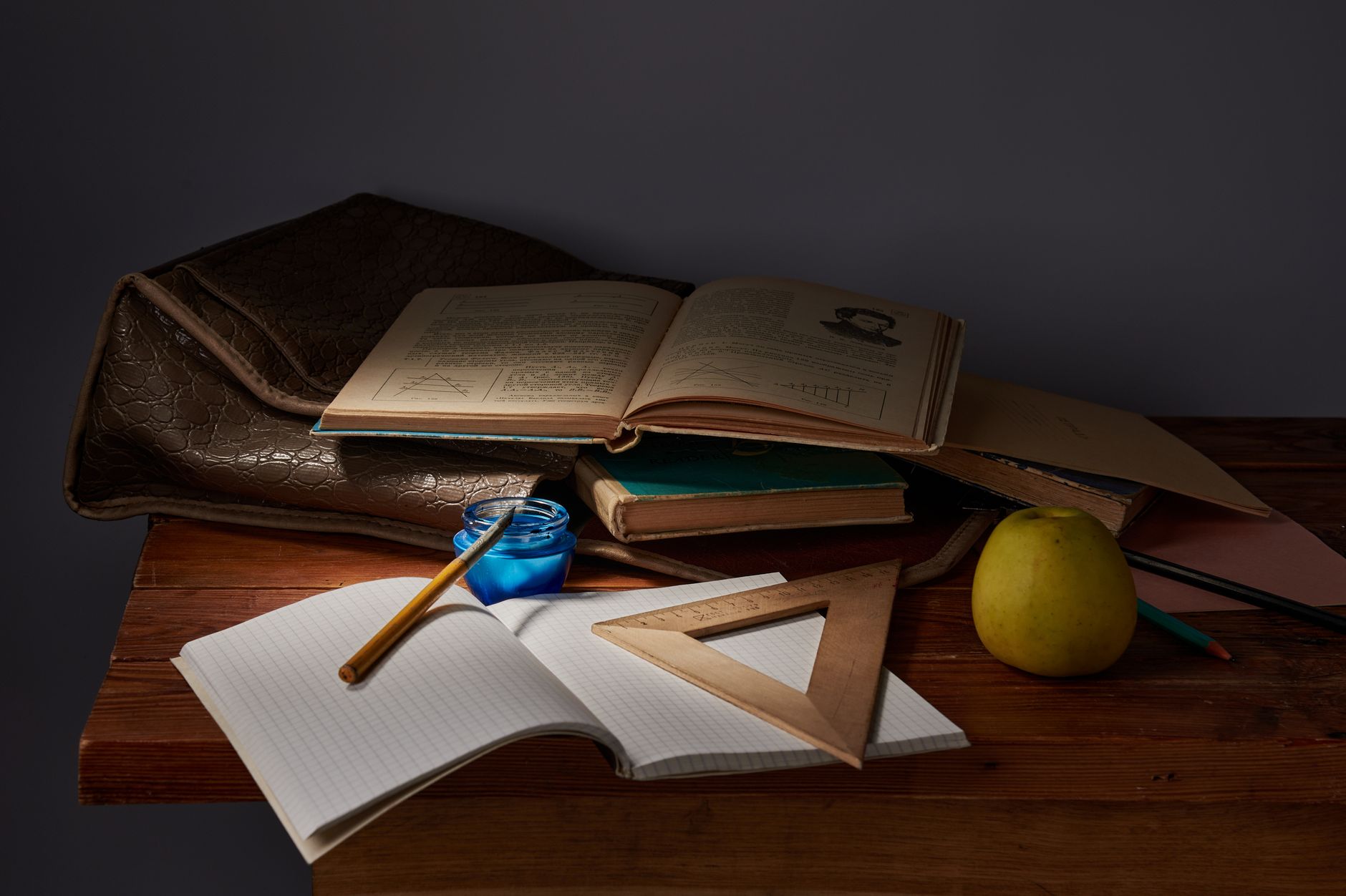 A classic still life composition featuring study materials, an apple, and a briefcase on a wooden table.