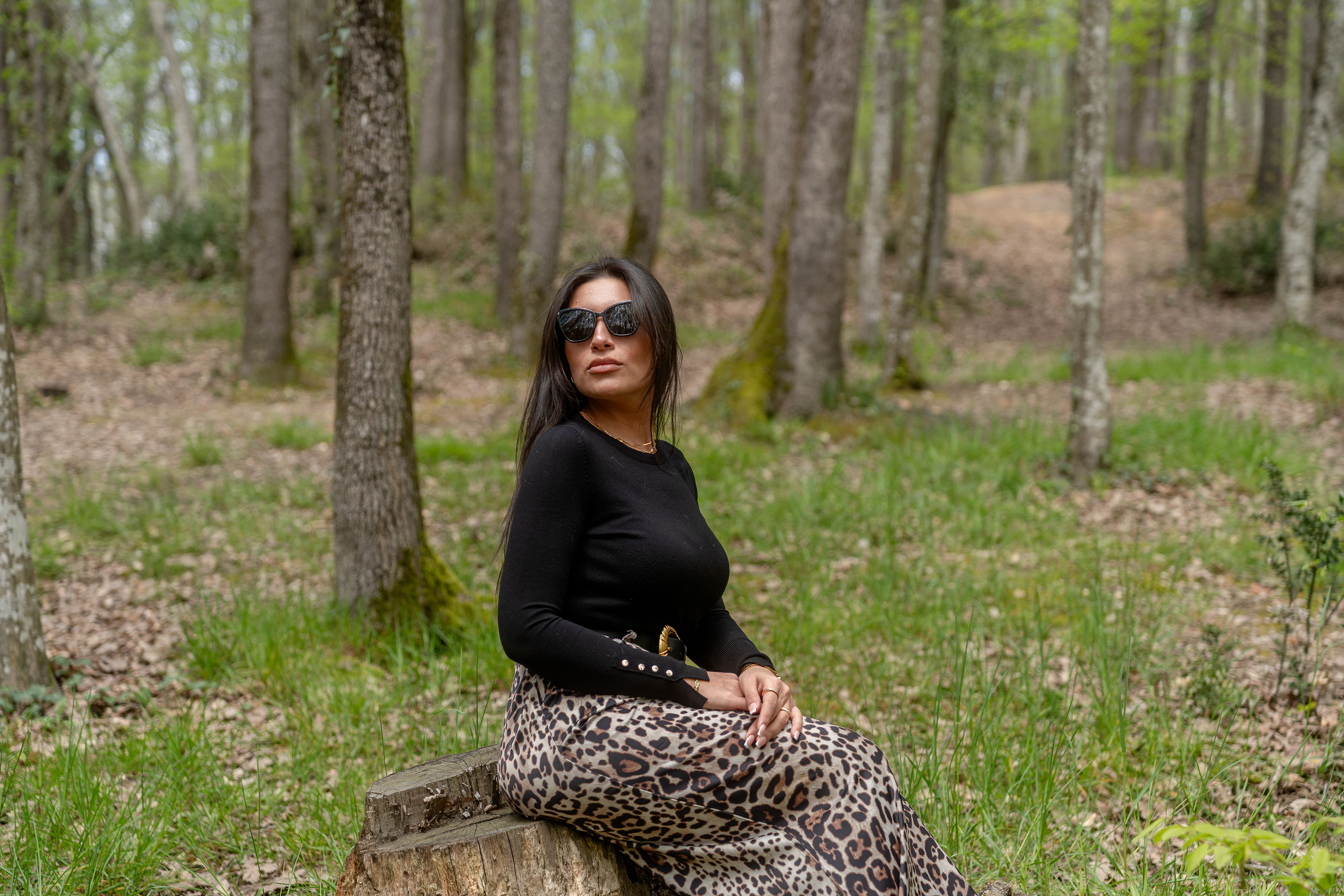 Stylish woman in sunglasses sitting on a log in a tranquil forest setting.