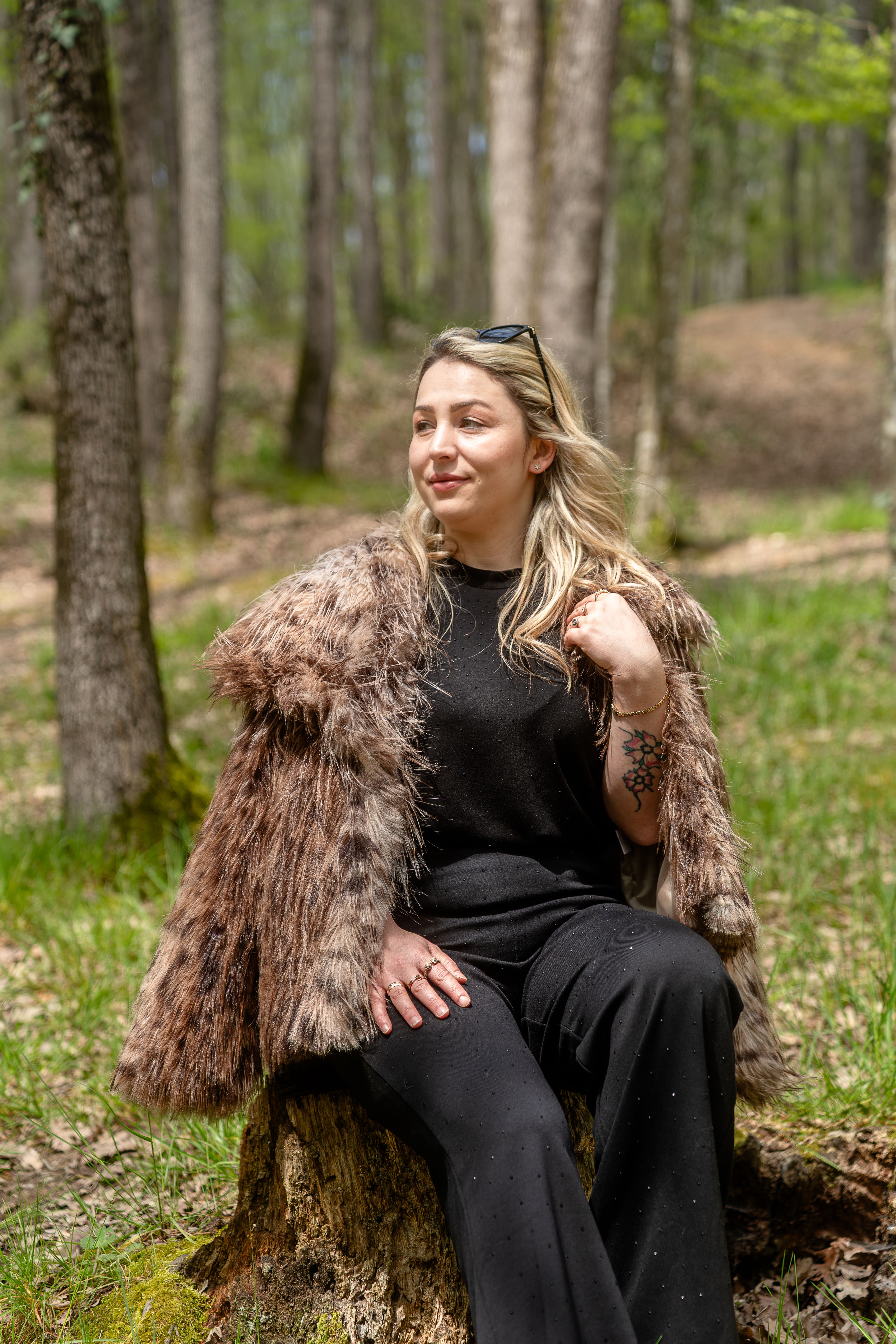 A woman sits on a tree stump in a forest, wearing a faux fur coat and enjoying nature. Warm lighting.