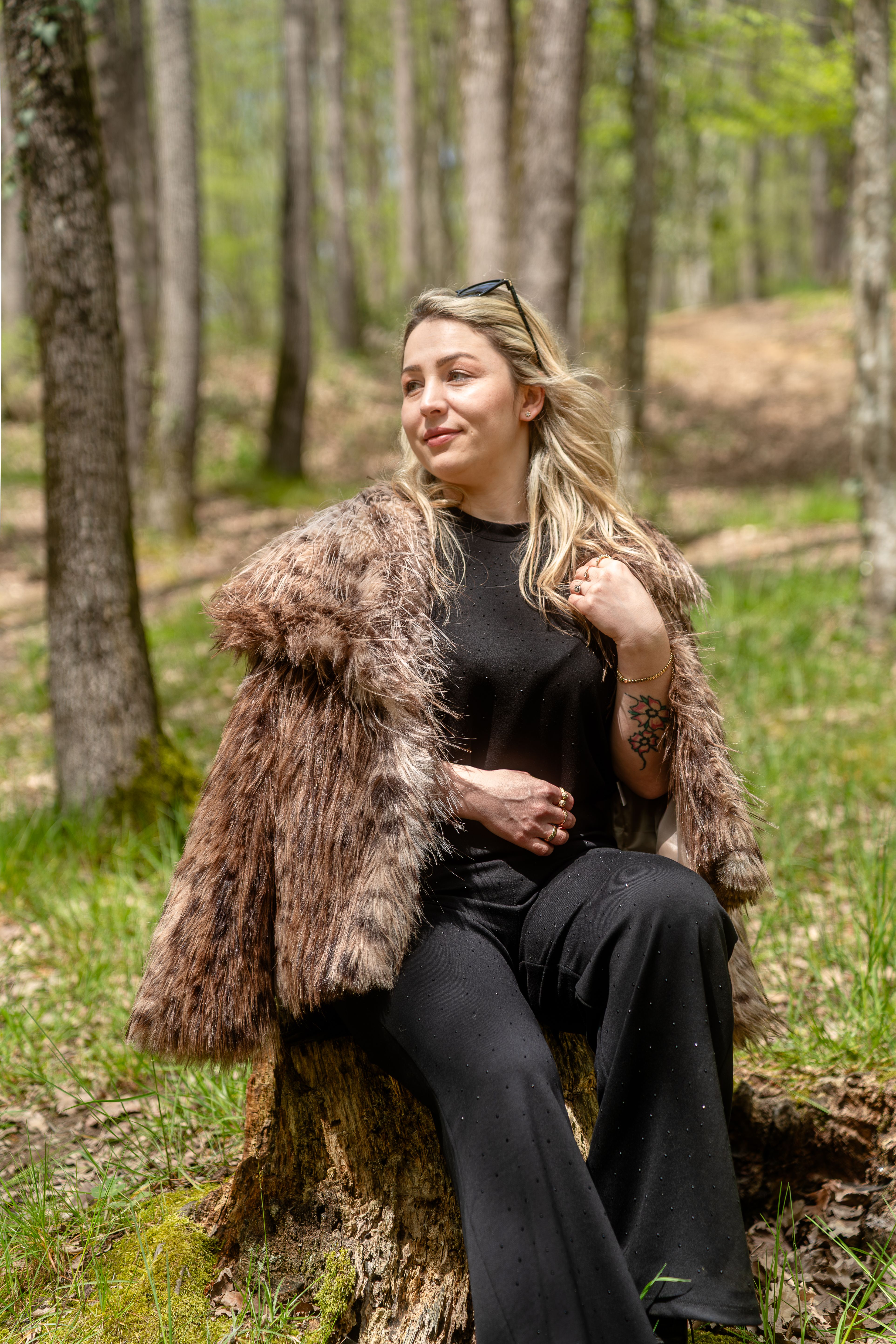 Woman in fur coat sitting on a tree stump in a serene forest setting, embodying natural elegance.