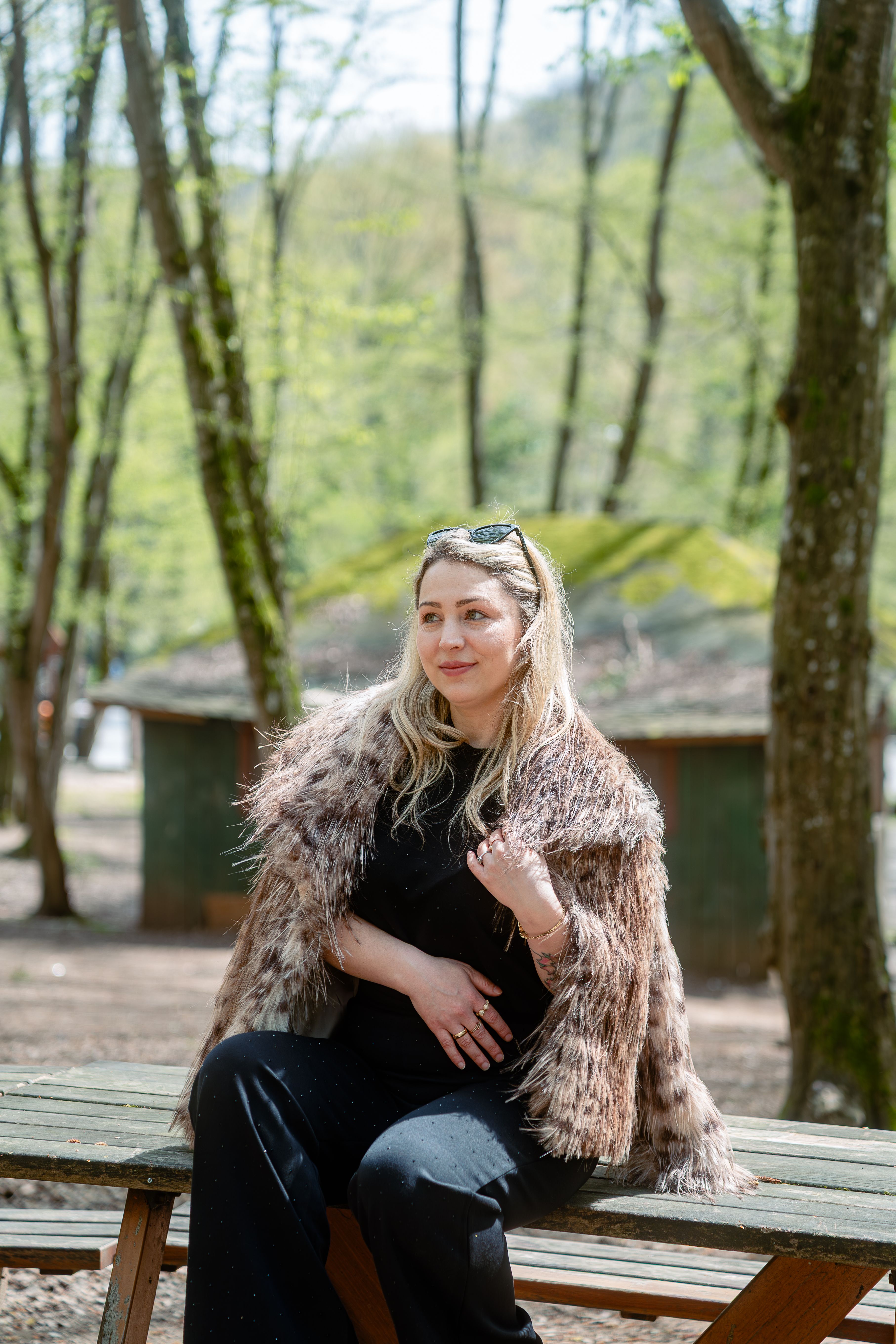 A stylish woman in faux fur sitting at a picnic table in a spring forest setting.