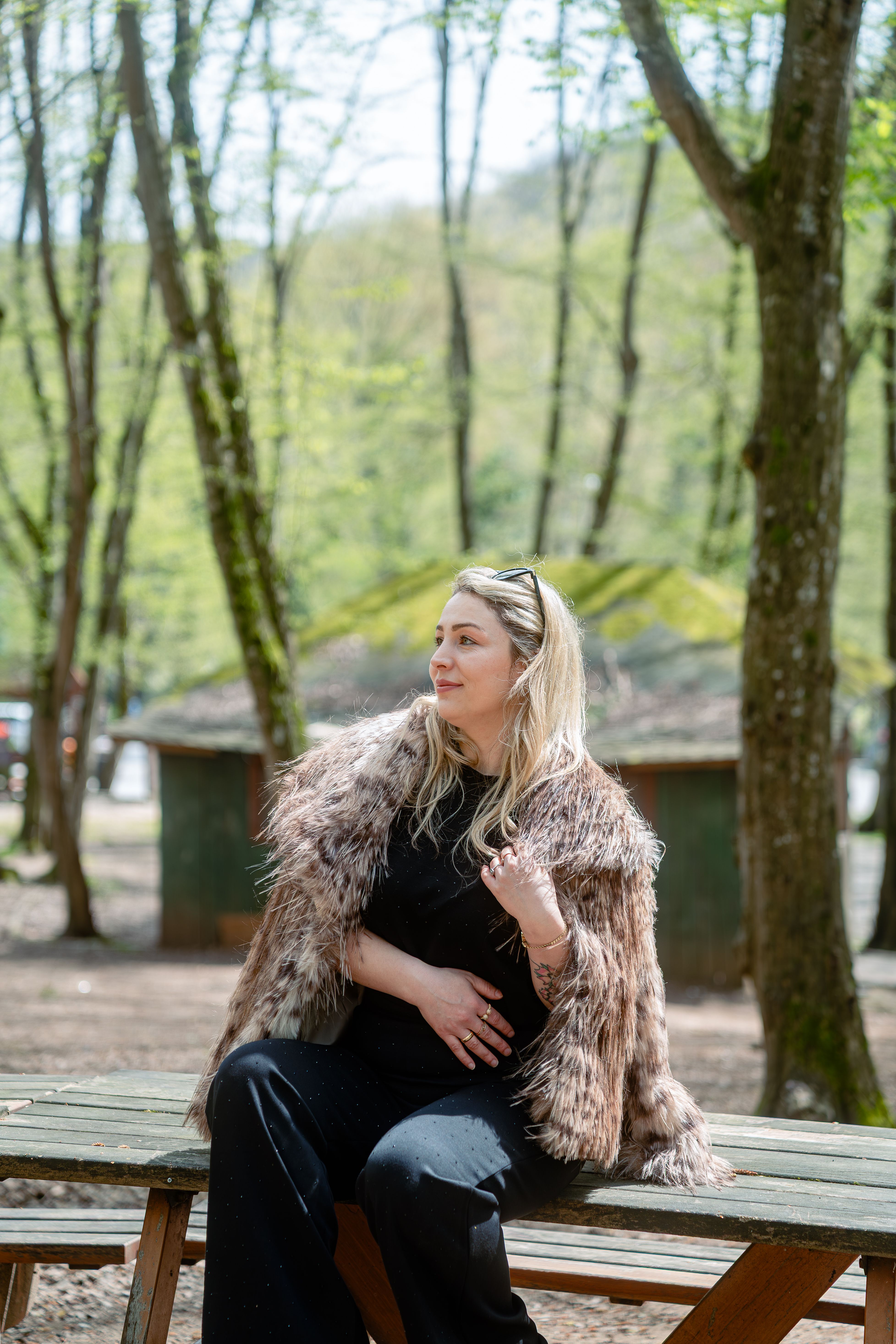 A fashionable woman wearing a fur coat sits at a picnic table in a serene forest setting.