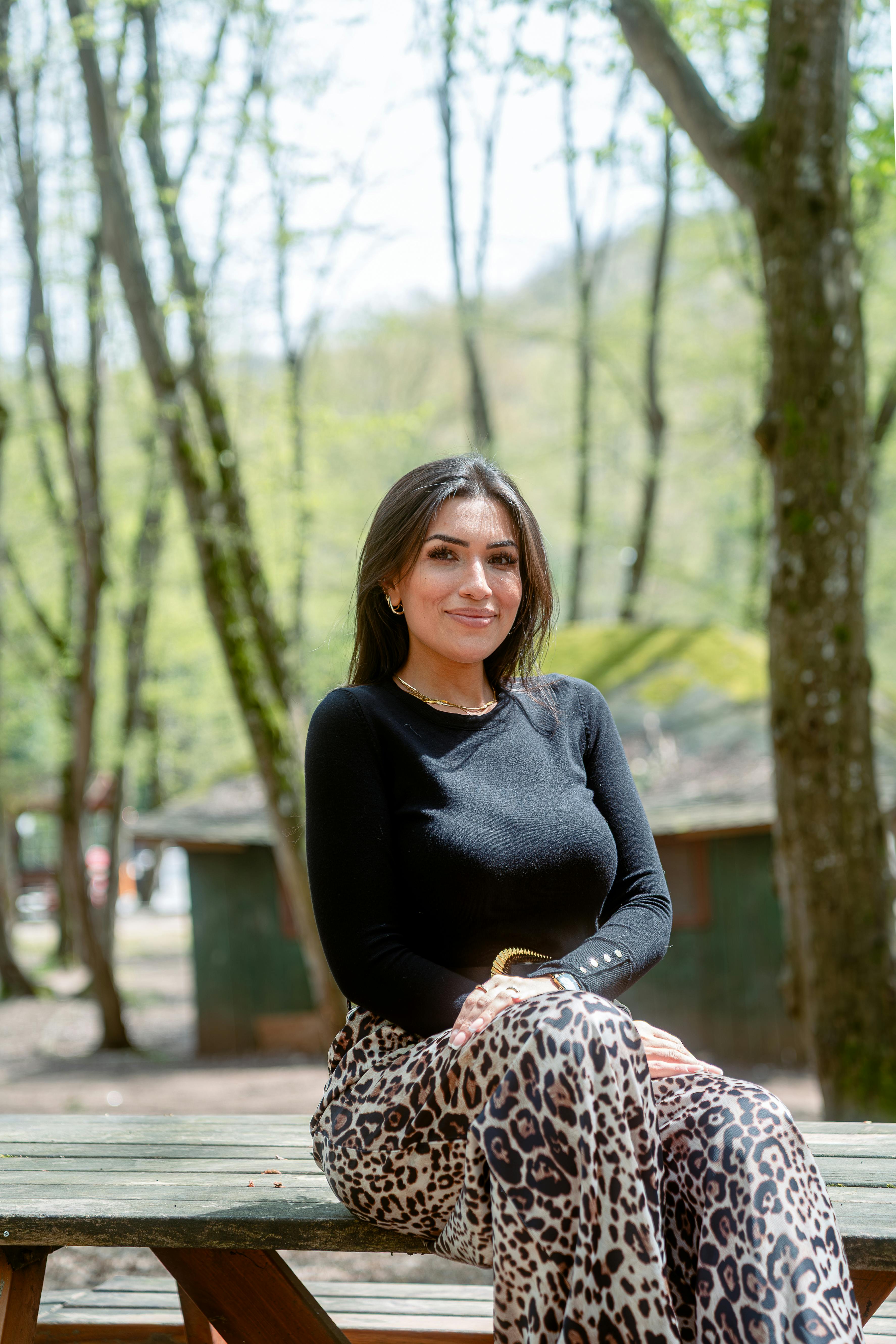 A woman sitting outdoors wearing a black top and leopard print skirt, surrounded by trees.