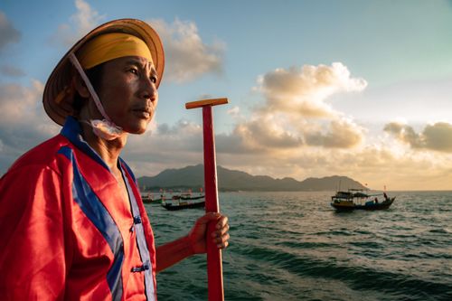 Free A traditional Vietnamese fisherman in colorful attire navigates at sunset over the sea with fishing boats in the background. Stock Photo