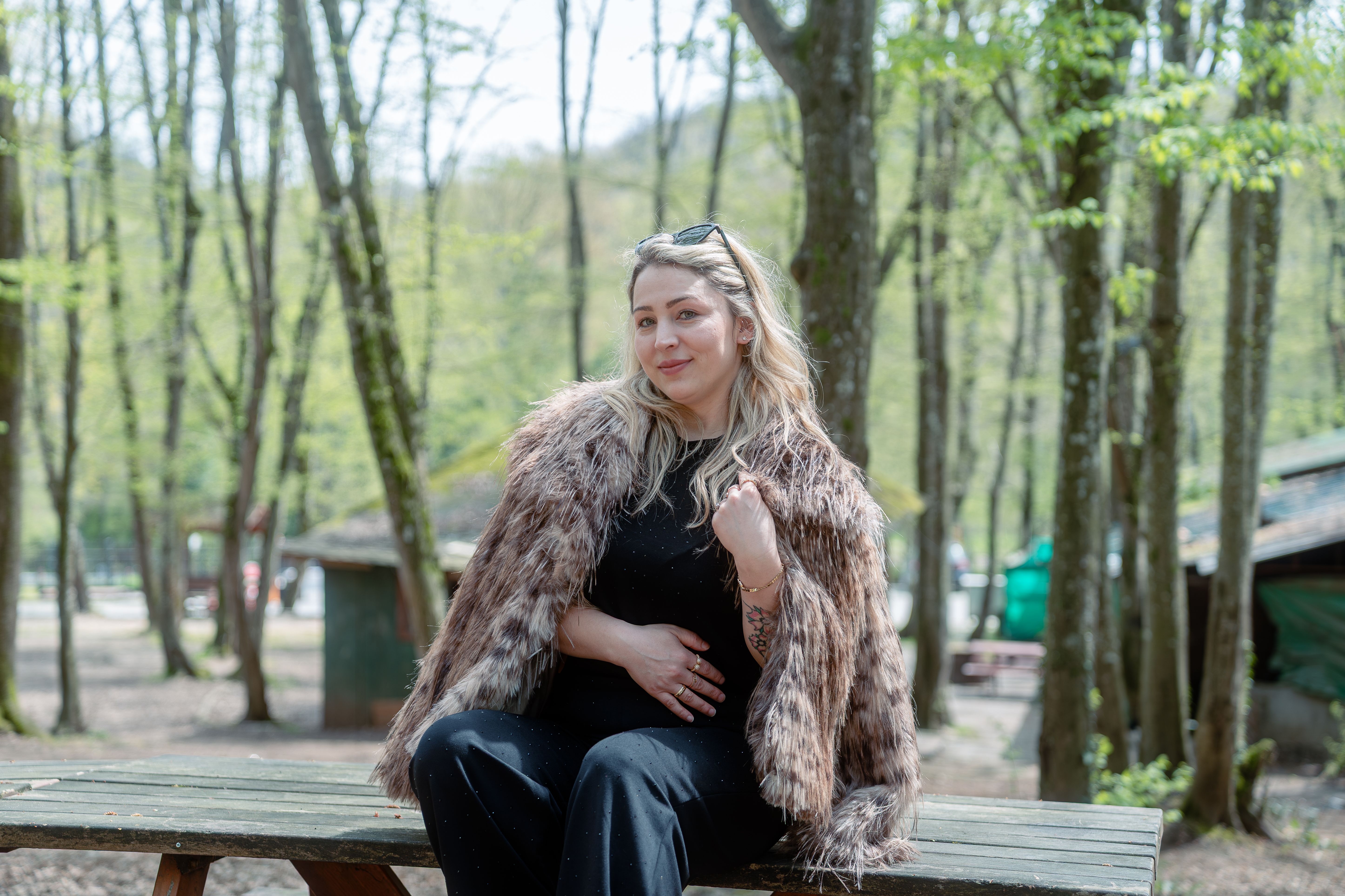 A fashionable woman sitting on a picnic table in a forest, wearing a fur coat.