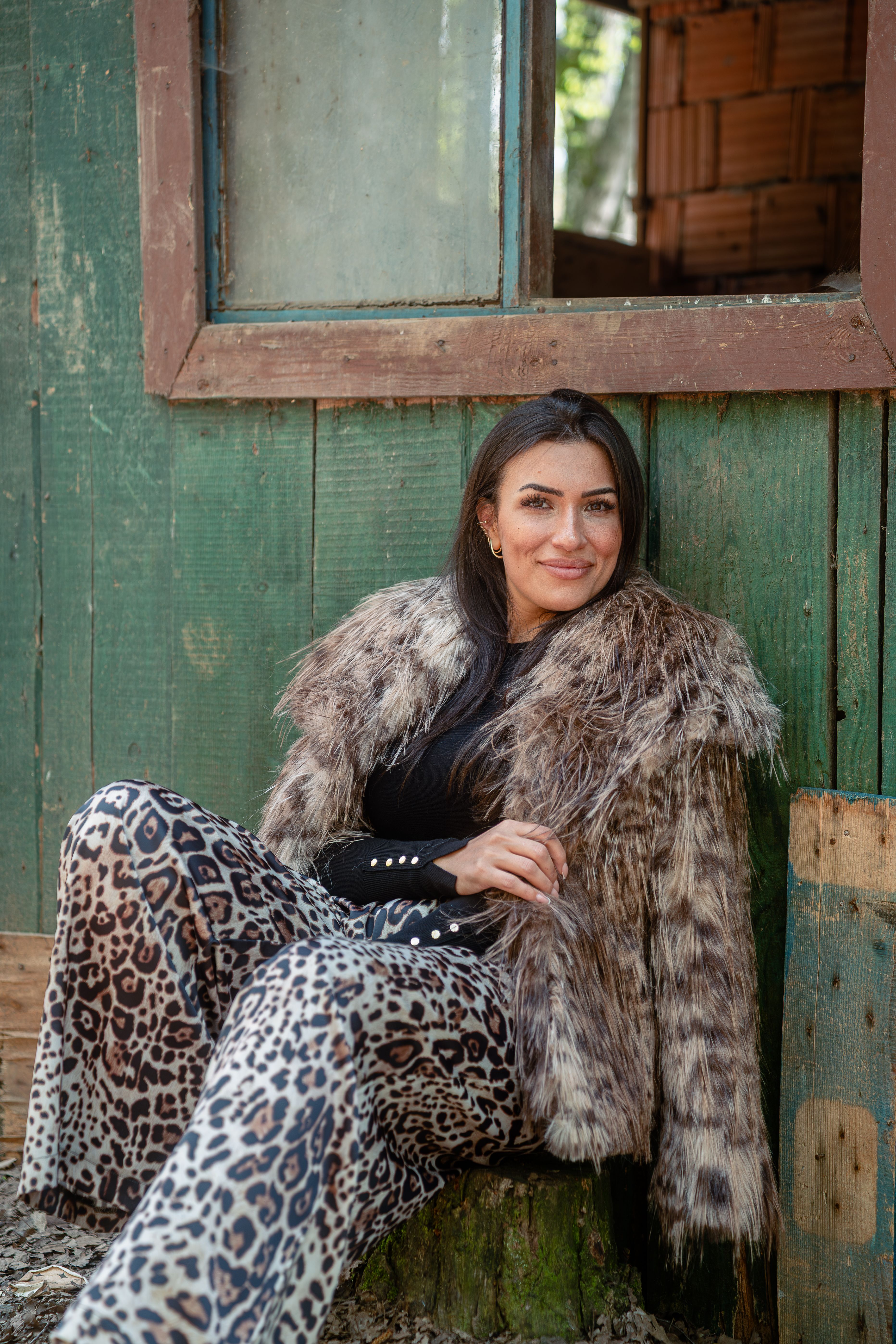 Stylish woman in a faux fur coat and leopard print pants sitting outdoors.