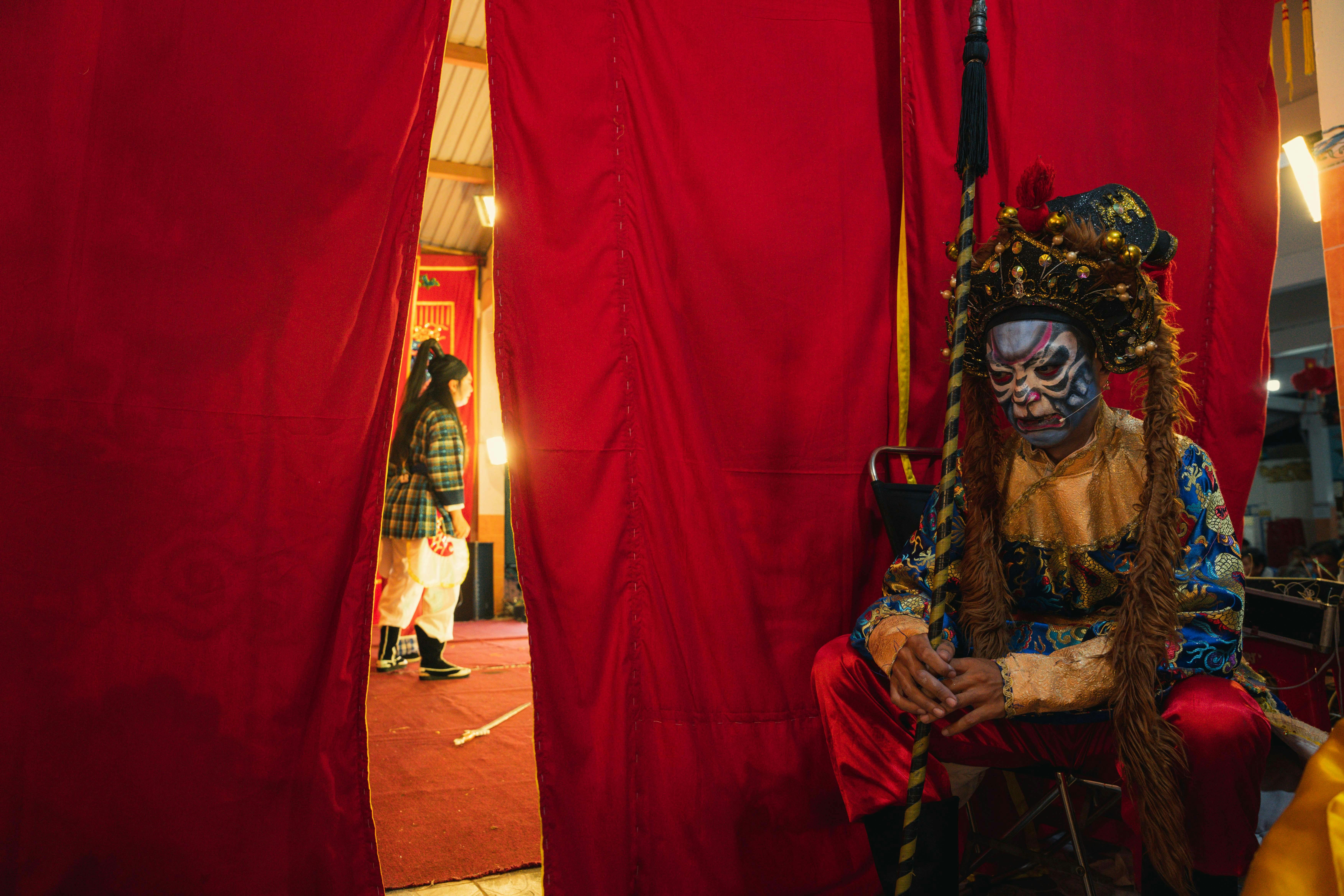 A Chinese opera performer in traditional costume and makeup awaits backstage in a vibrant setting.