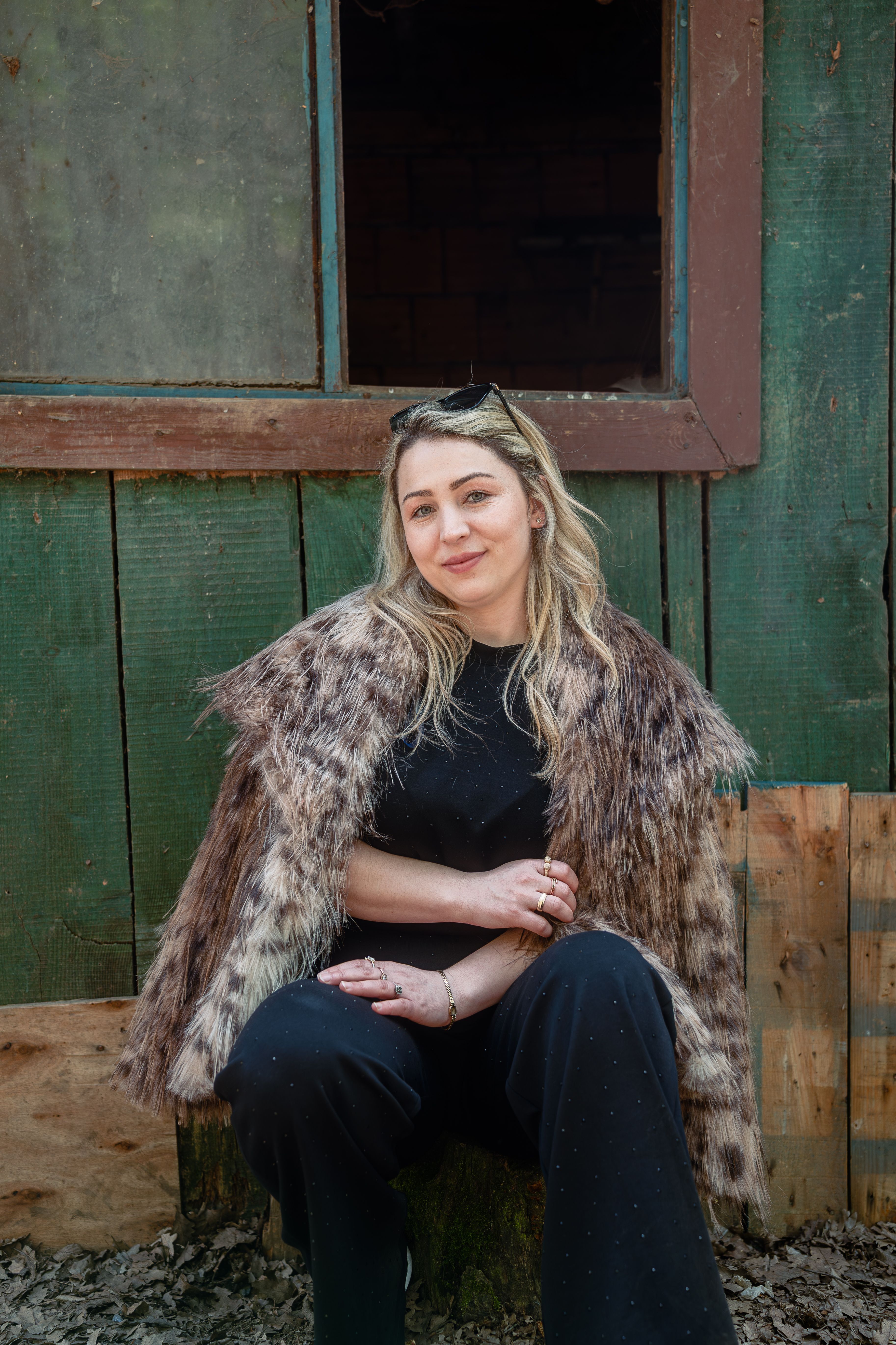 Stylish woman wearing a faux fur coat sits confidently outside a weathered wooden building.